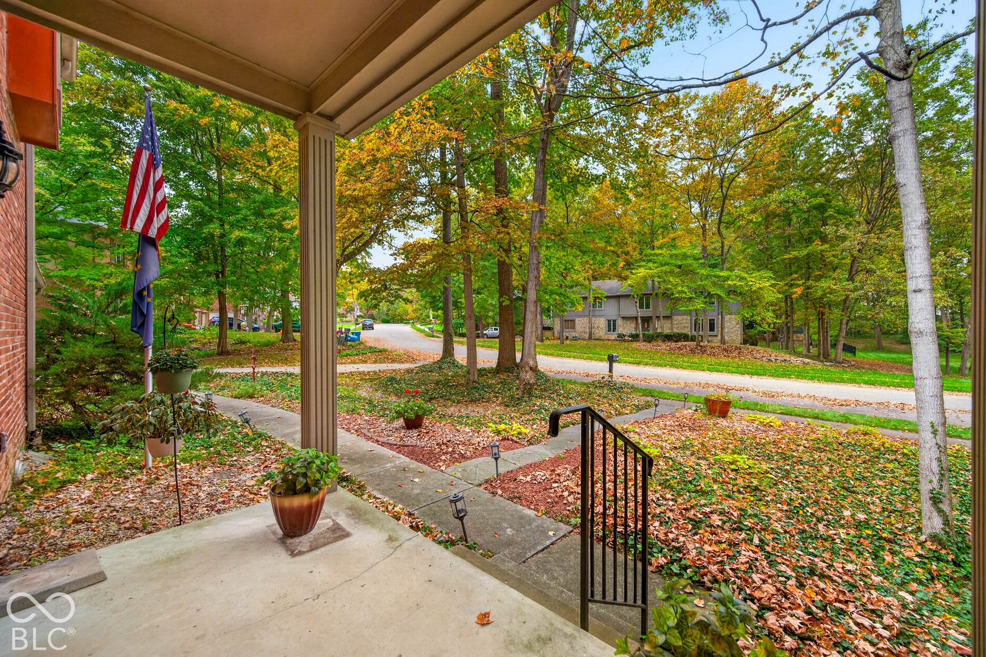 Covered front porch featuring a concrete floor, support columns, and metal handrails, overlooking a lot with mature trees