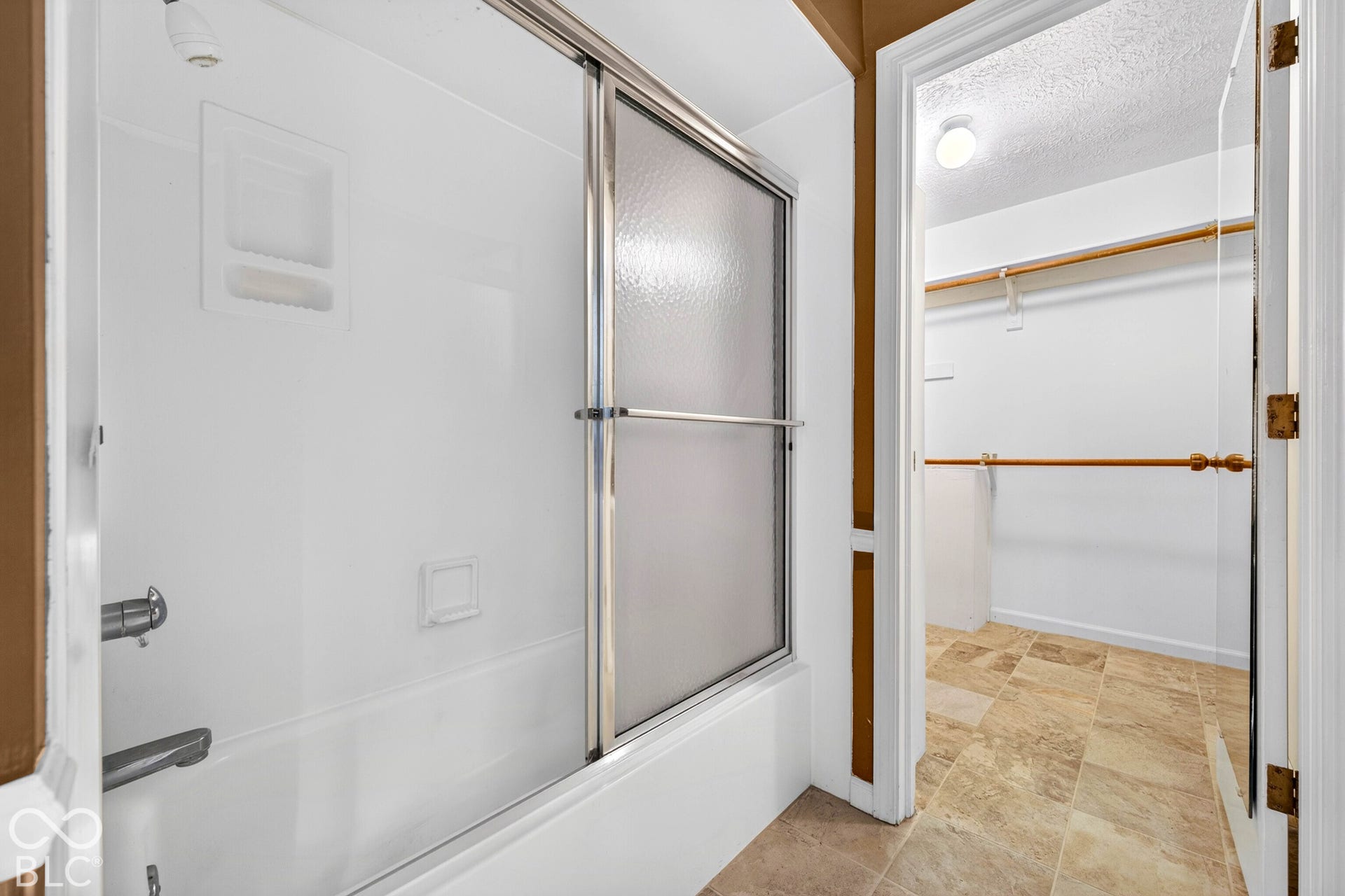 Shower with a sliding glass door and integrated shelf, paired with a walk-in closet featuring built-in wooden shelving and stone-finish flooring