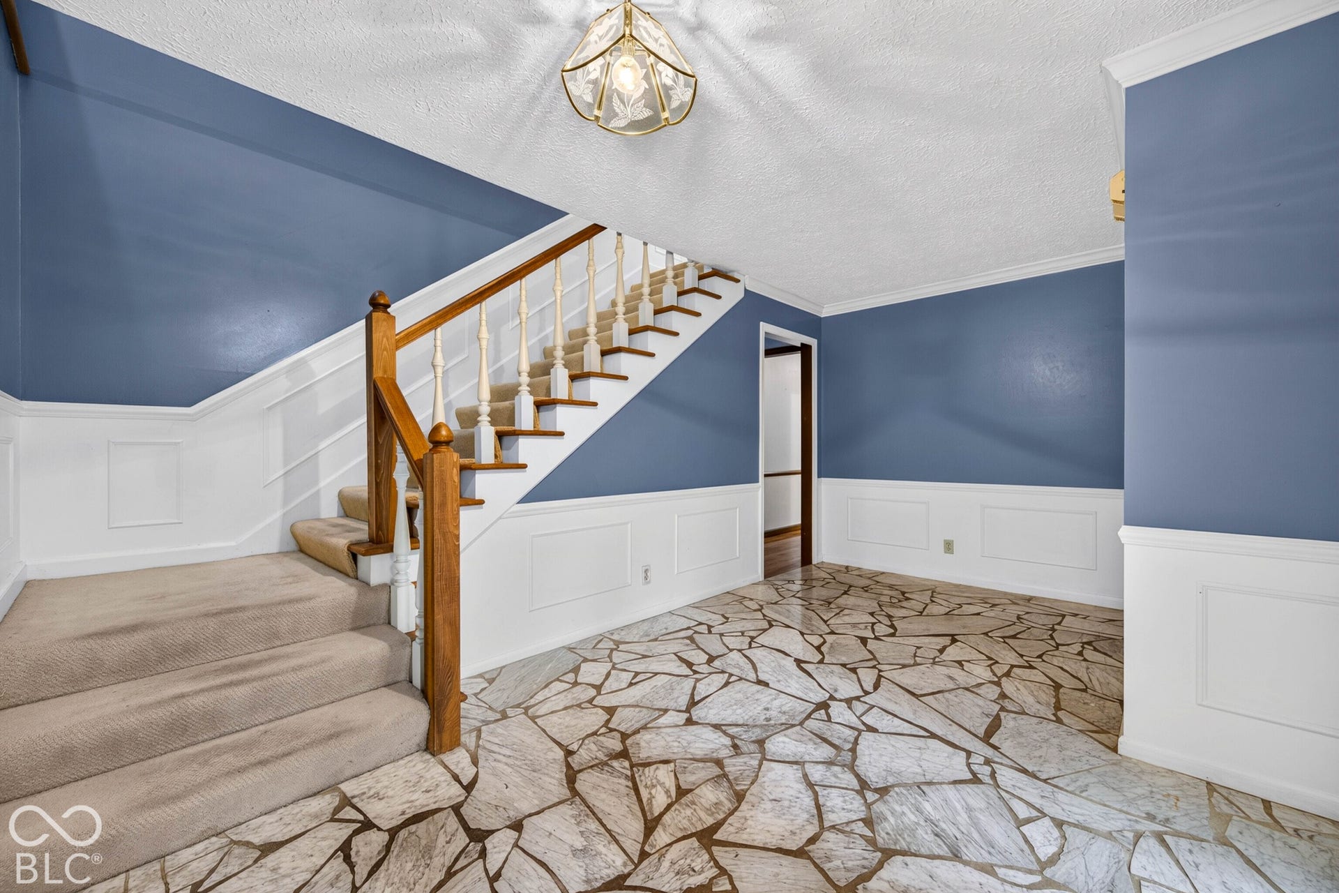 Foyer featuring stone flooring with inlaid grout, white wainscoting, a wooden staircase railing with decorative balusters, and a glass-paneled ceiling fixture