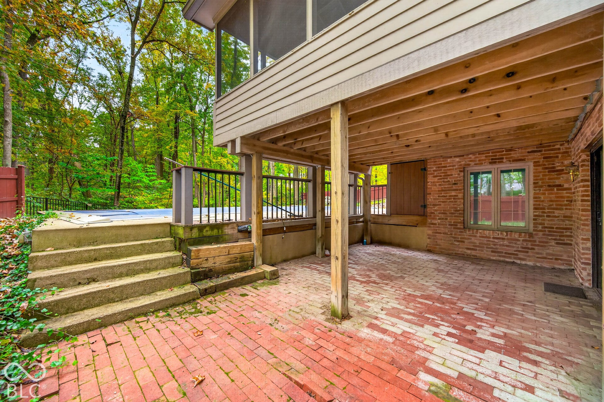 Covered patio featuring red brick paving, exposed ceiling joists, and exterior brick walls, with concrete steps leading to an elevated outdoor area enclosed by metal railings