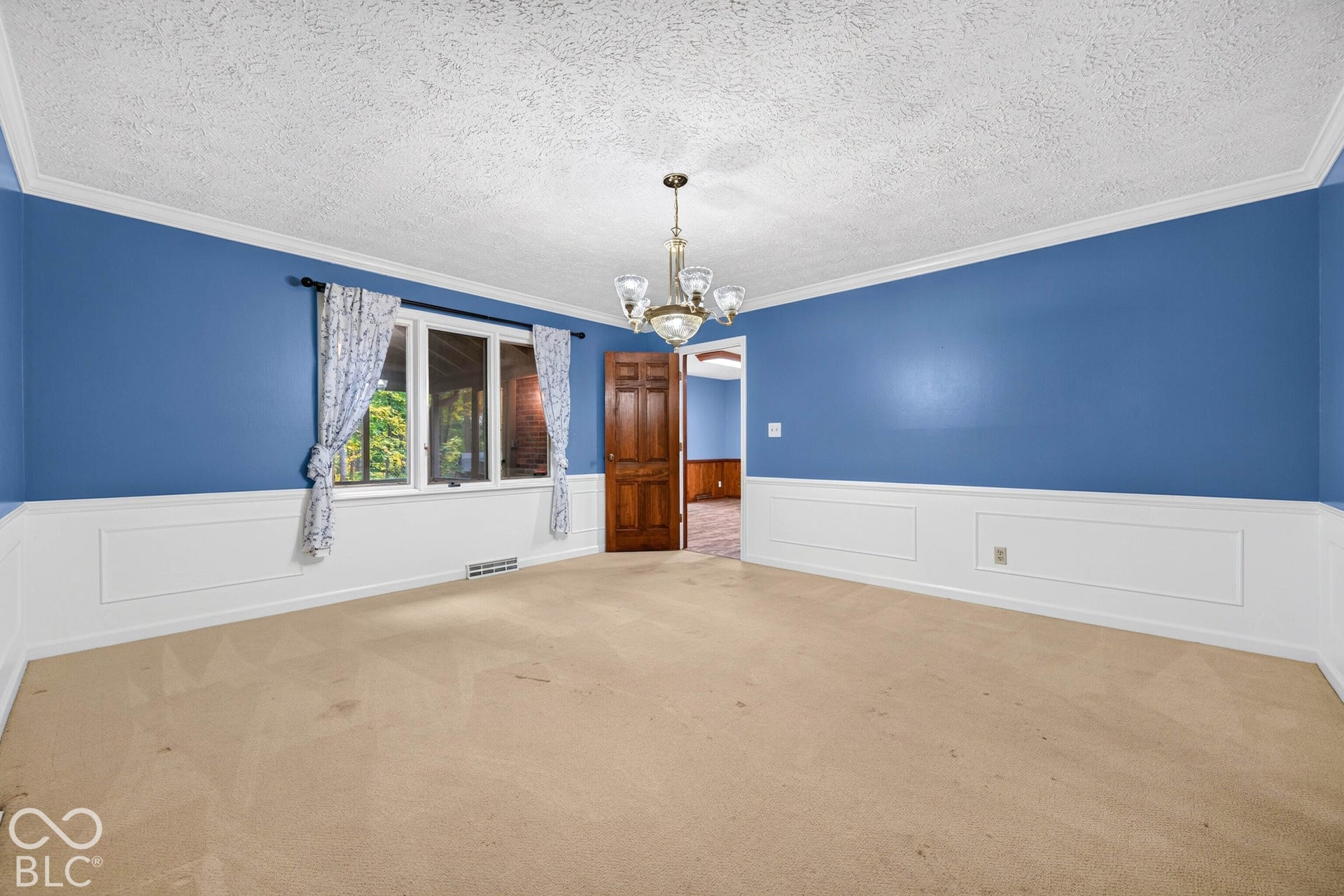 Room featuring crown molding, lower wall paneling, multi-pane window, wood-finish interior door, and a multi-arm chandelier