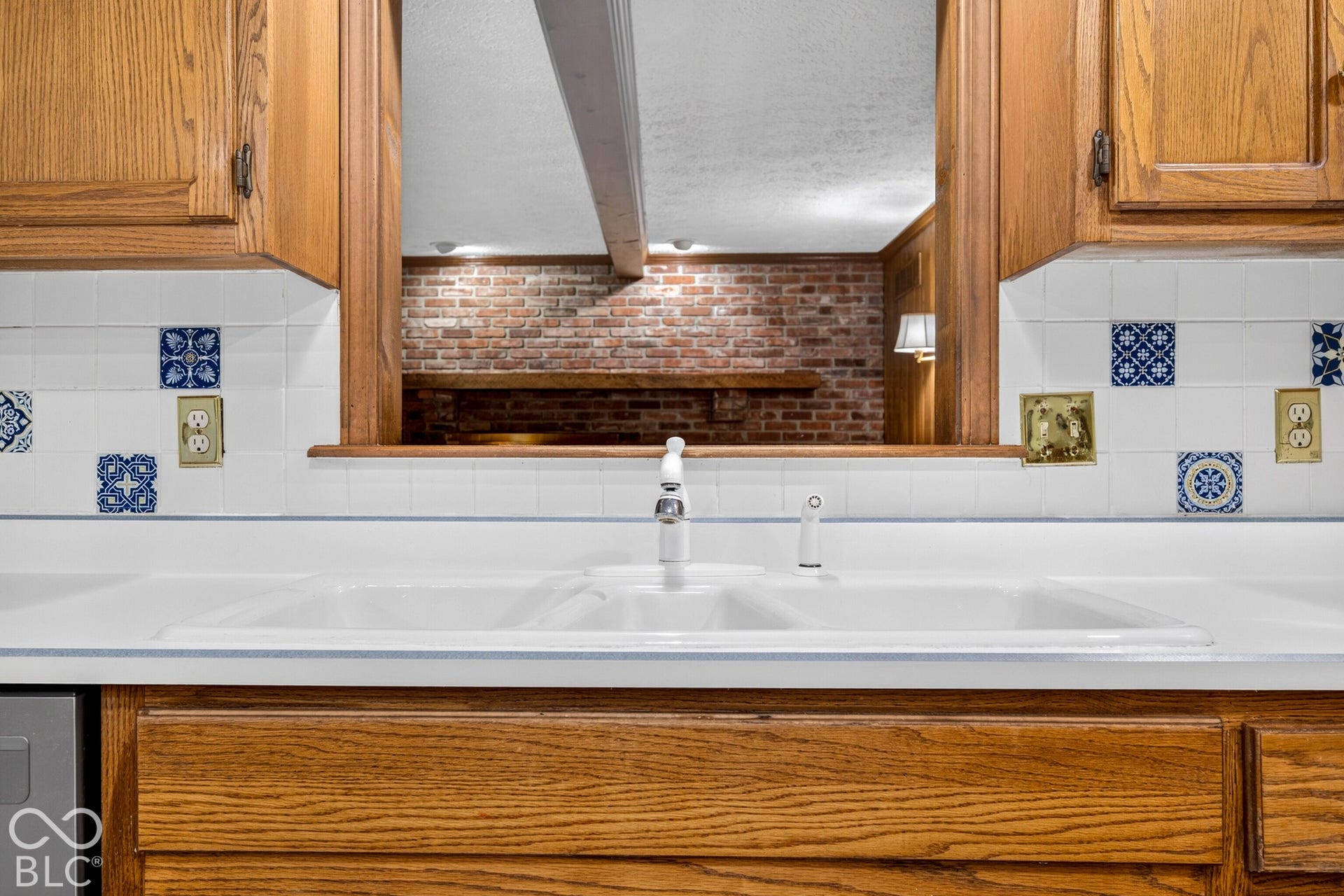 Kitchen features a white double-basin sink, wood-finish cabinetry, decorative accent tiling, and a pass-through opening framed by exposed wood trim