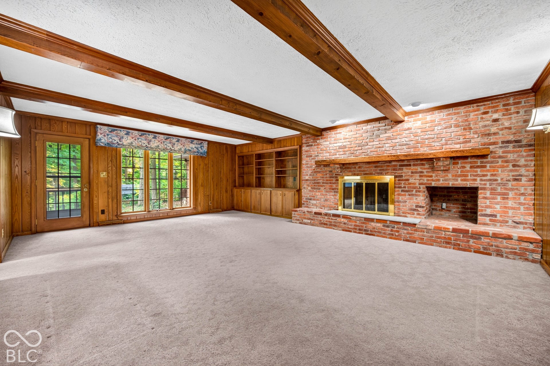 Expansive room featuring a floor-to-ceiling red brick fireplace with a wood mantel, wood beam ceiling details, wood-paneled walls, and built-in shelving