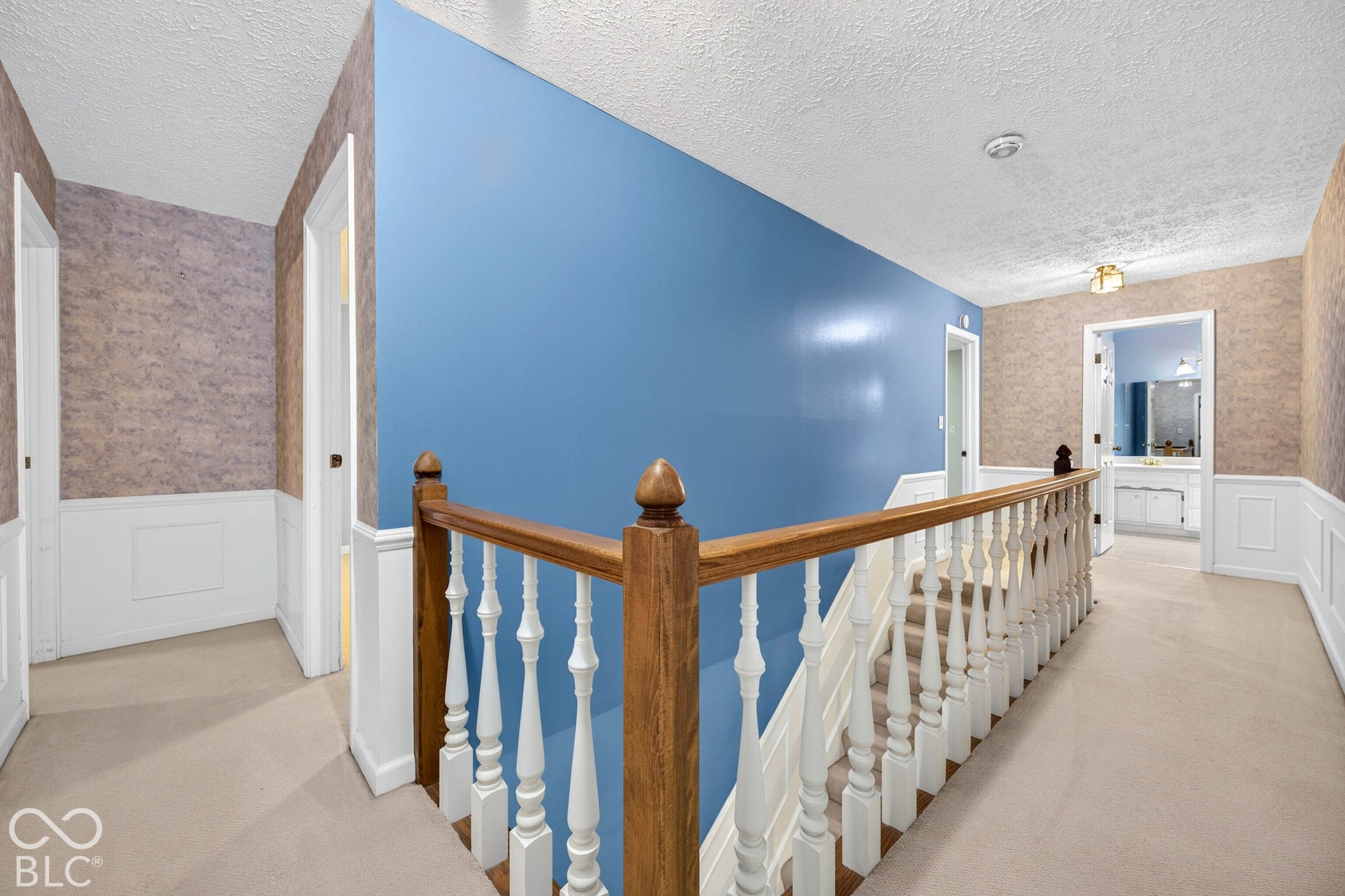 Hallway featuring wood-finish banister, white spindle railing, wainscoting, and textured wallpaper