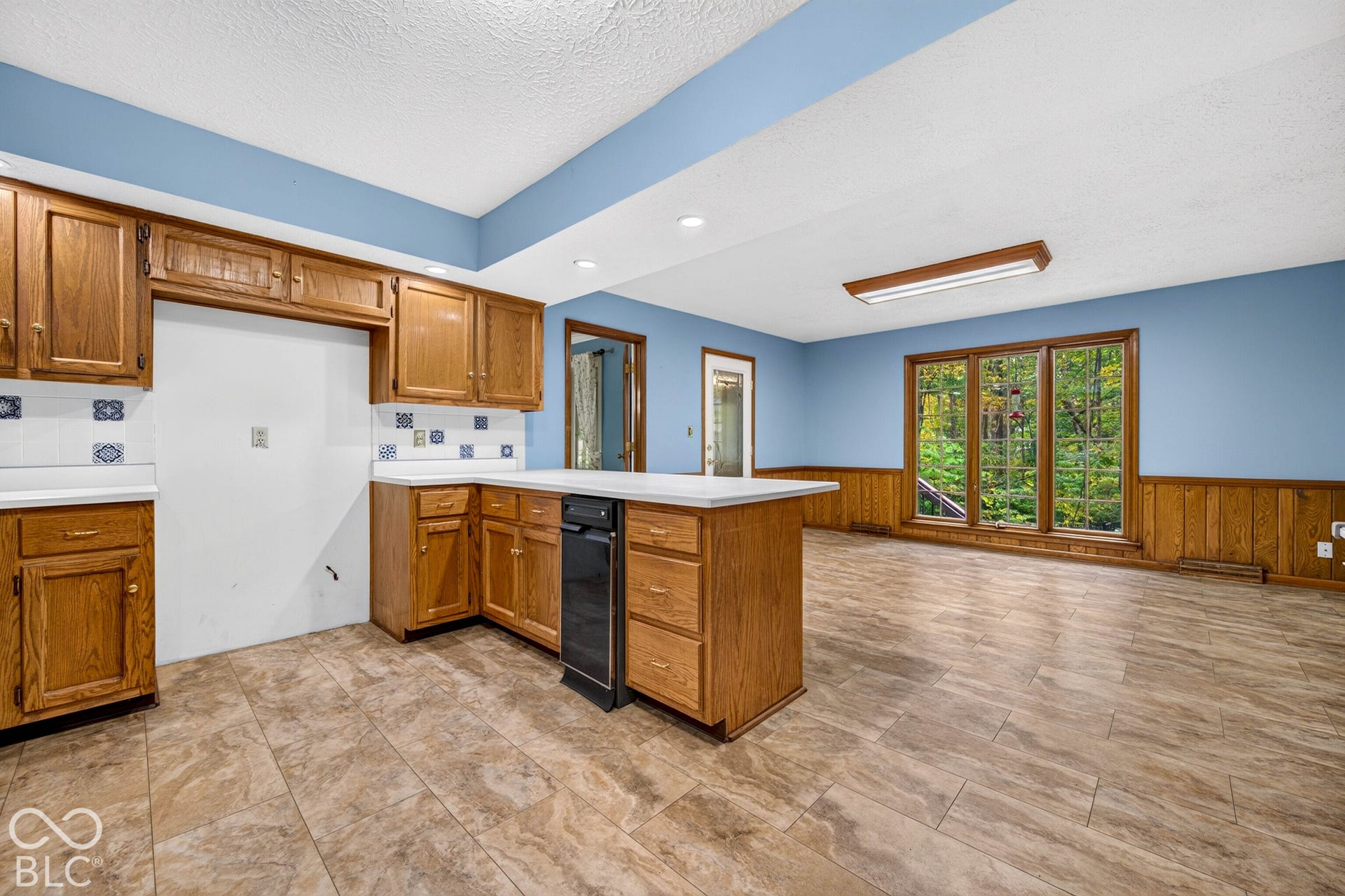 Kitchen featuring medium-tone wood cabinetry, solid-surface countertops, tile flooring, wood wainscoting, and a triple-pane window