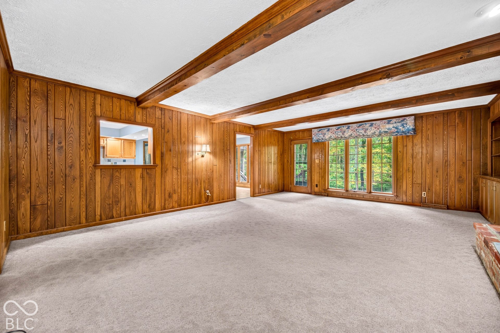 Wood-paneled interior featuring exposed wooden ceiling beams, built-in cabinetry, large windows with natural scenery, and a brick hearth