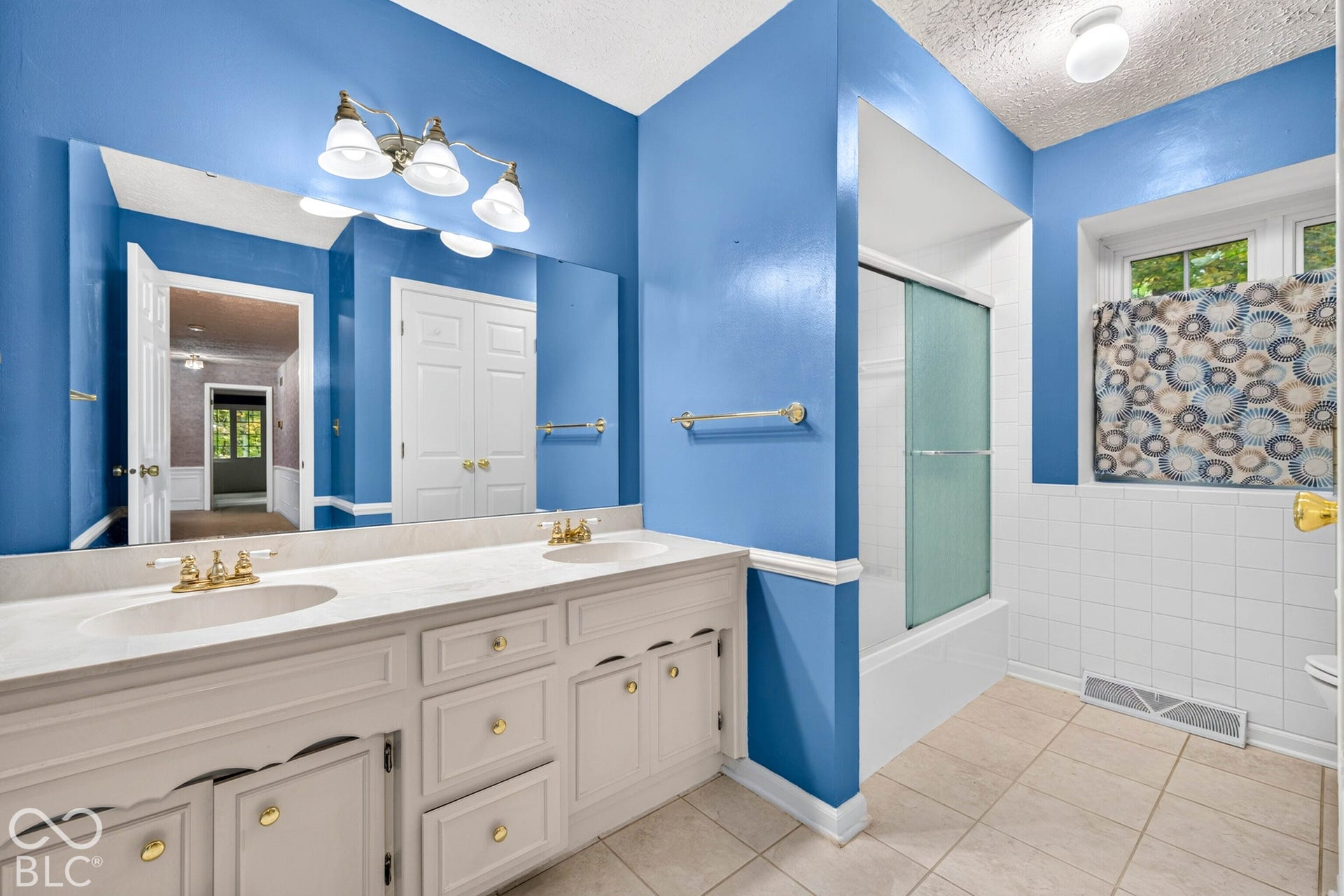 Dual-sink vanity with a solid-surface countertop, full-width vanity mirror, brass-finish fixtures, and a shower-tub combination with sliding glass doors