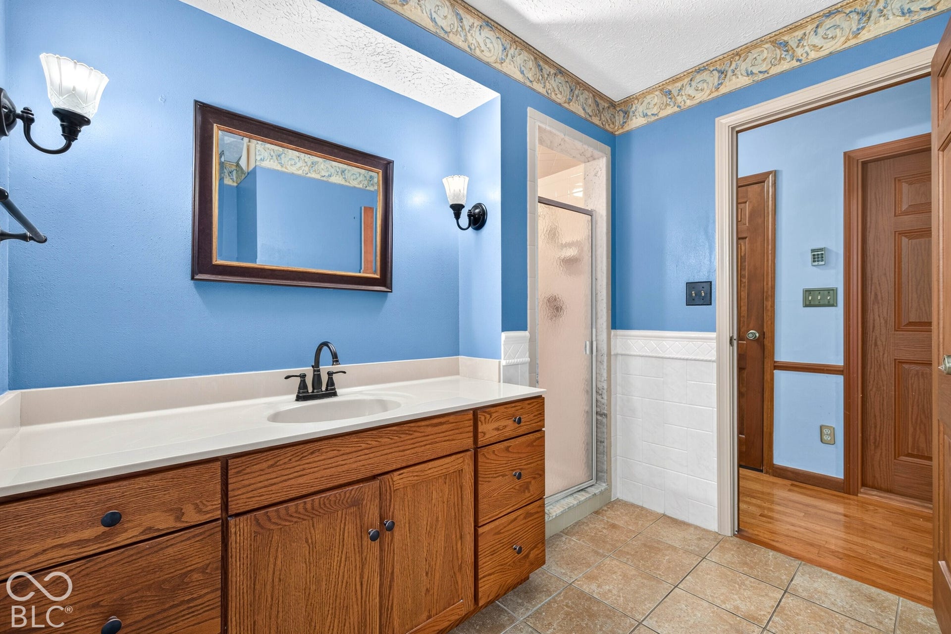 Bathroom featuring a wooden vanity with integrated sink, stone-finish countertop, dual wall-mounted light fixtures, white wainscoting, and a walk-in shower with a glass door