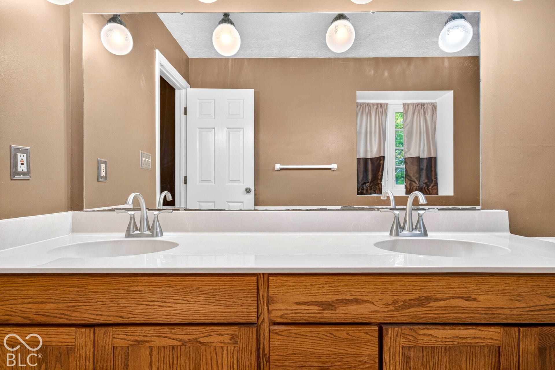 Dual-sink vanity featuring a white countertop with integrated basins, wood-finish cabinetry, brushed nickel faucets, and a full-width wall mirror with integrated lighting