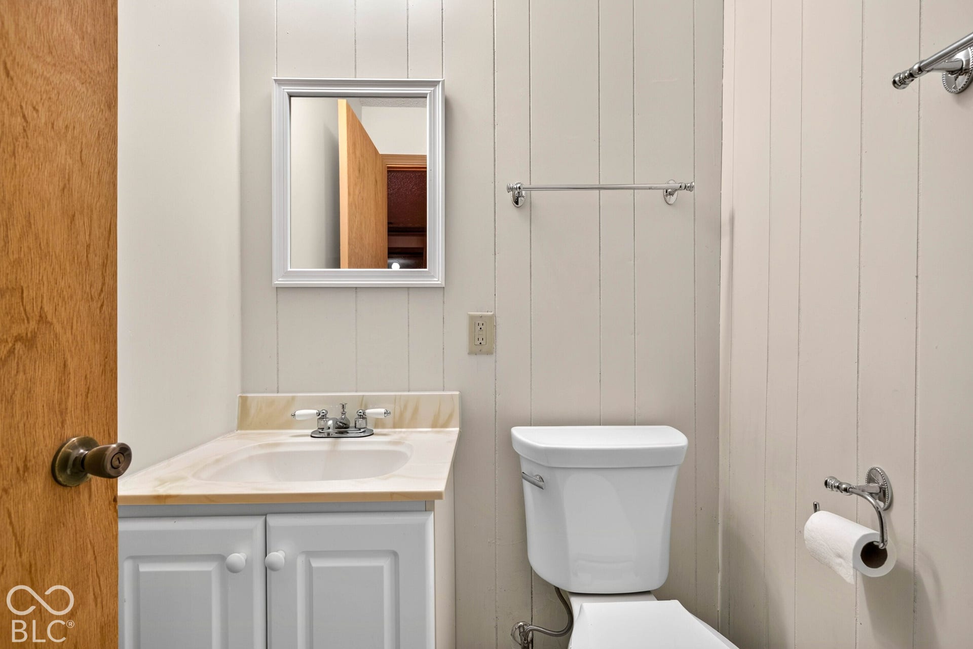 Bathroom featuring vertical plank wall paneling, a vanity with a light-toned countertop, an integrated sink with a dual-handle faucet, a framed mirror, and chrome hardware