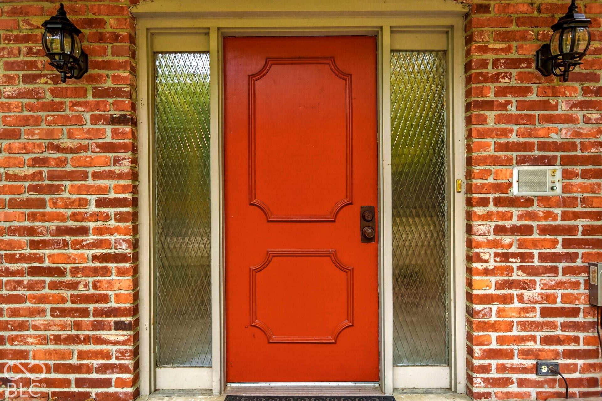 Exterior entryway featuring a red panel door, dual sidelights with diamond-patterned security glass, and red brick masonry facade accented by two dark-finish wall-mounted lanterns