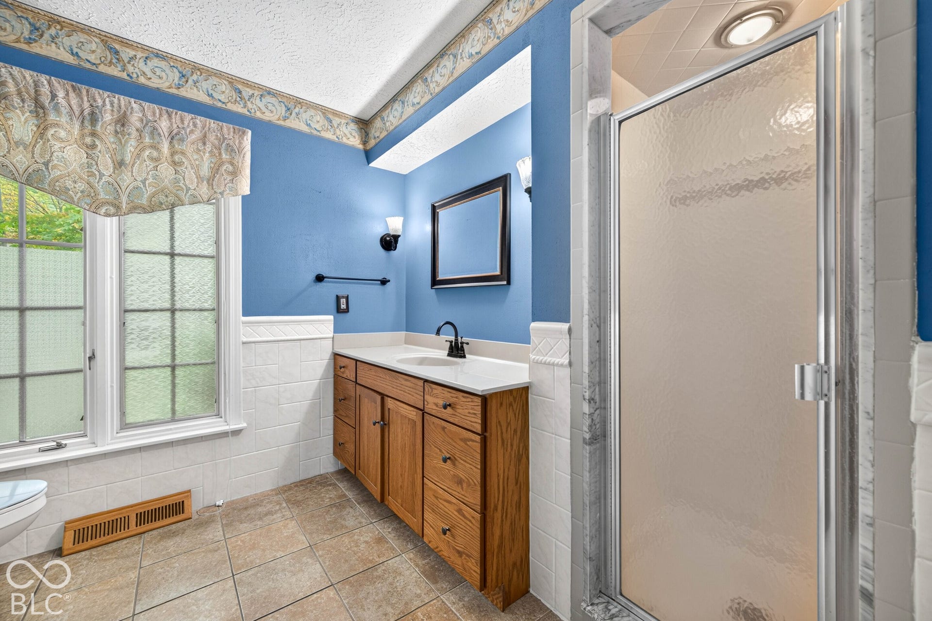 Wood-finish cabinetry with a white countertop, tiled shower enclosure with a glass door, ceramic tile flooring, and wall-mounted light fixtures
