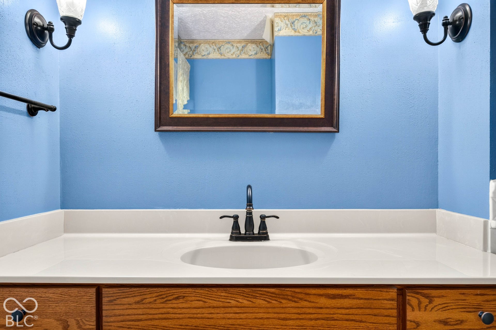 Vanity featuring a white countertop with an integrated basin, wood-finish cabinetry, a dark-toned widespread faucet, a framed wall mirror, and two wall-mounted light fixtures