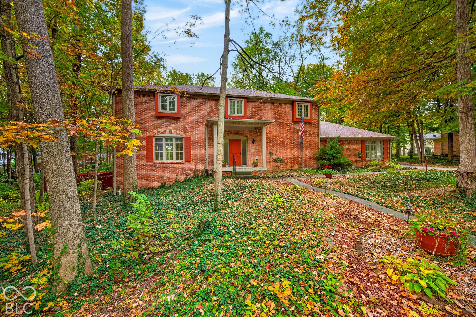 Two-story brick exterior featuring a covered front entryway, multi-pane windows with shutters, and a concrete walkway set within a wooded lot