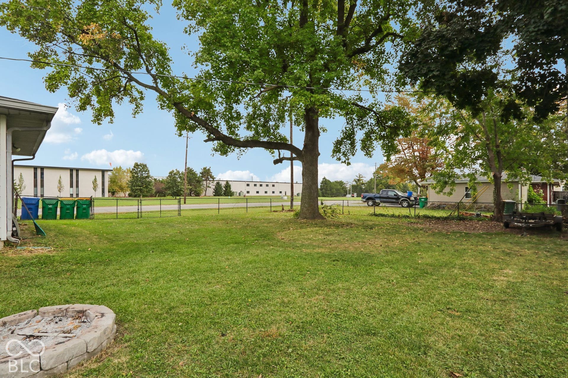 Expansive backyard featuring a mature canopy tree, stone-bordered fire pit, and chain-link perimeter fencing