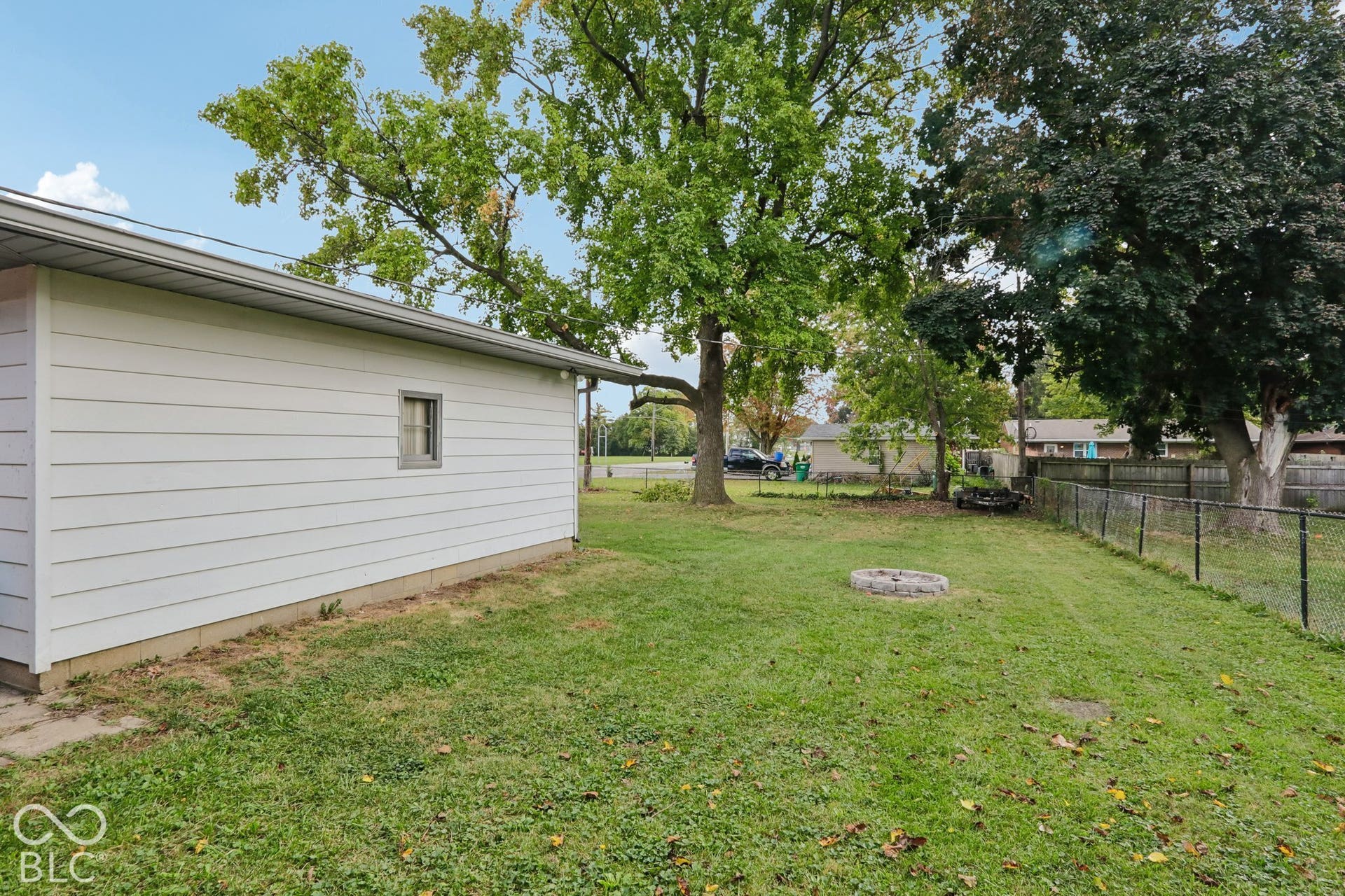 Backyard featuring horizontal vinyl siding, mature trees, chain-link fencing, and a stone-bordered fire pit