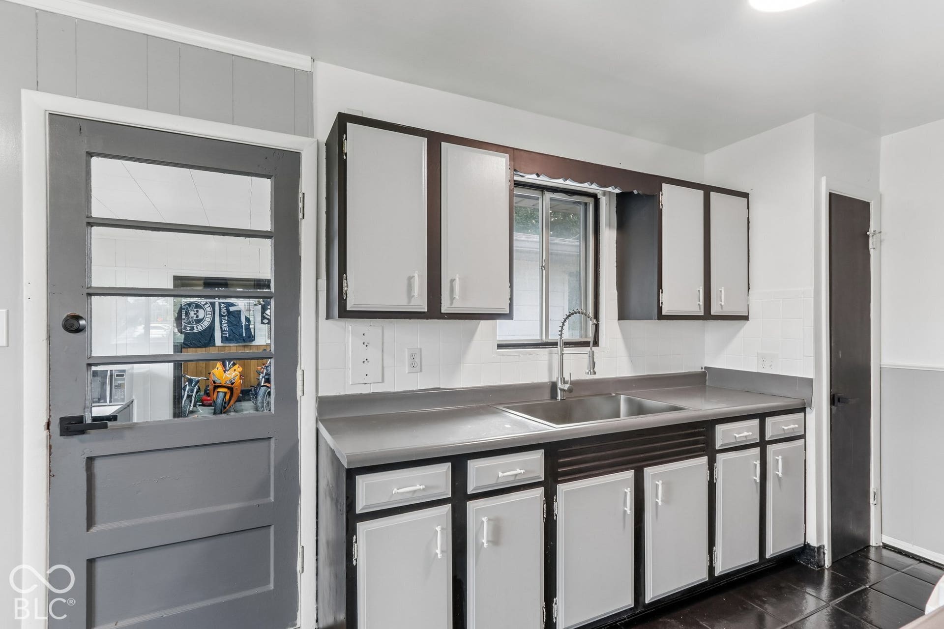 Kitchen featuring contrast-painted cabinetry, stainless steel sink with gooseneck faucet, solid-surface countertops, tile backsplash, and paneled exterior door with glass insert