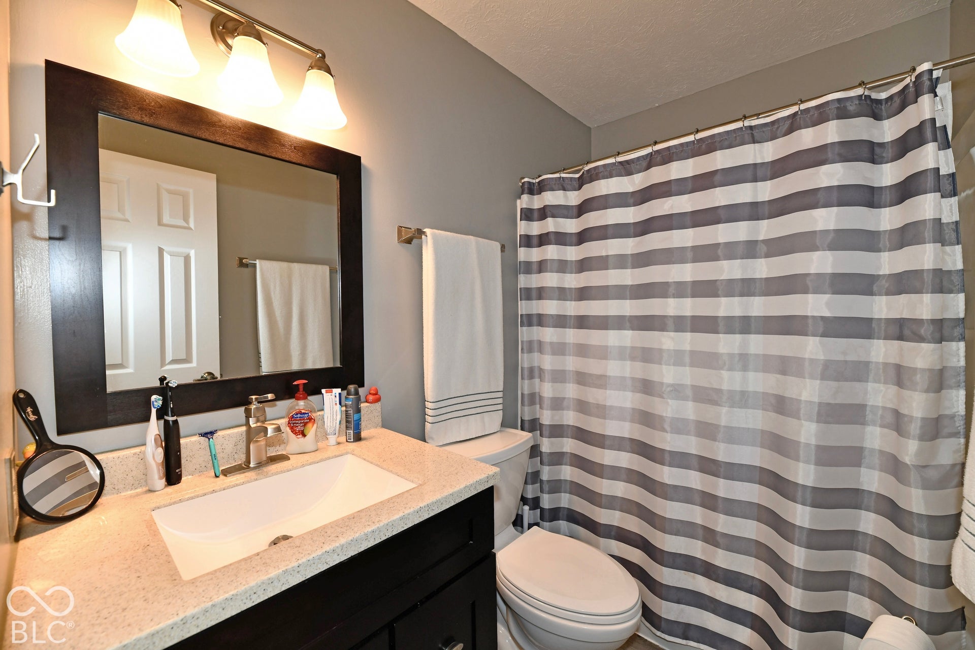Bathroom vanity featuring a dark-finish wood cabinet, light-toned stone-composite countertop, undermount sink, three-light fixture, and a large framed mirror