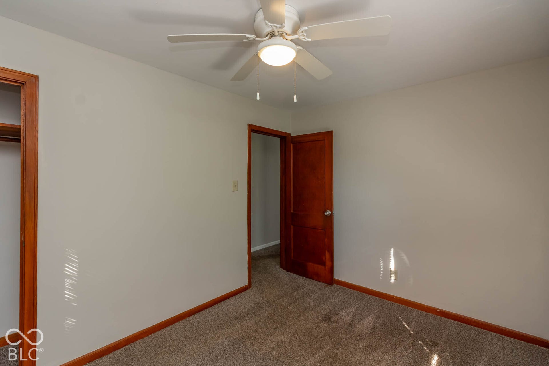 Room featuring wall-to-wall carpeting, wood-trim door frames, a solid-core wood door, and a multi-blade ceiling fan with integrated lighting