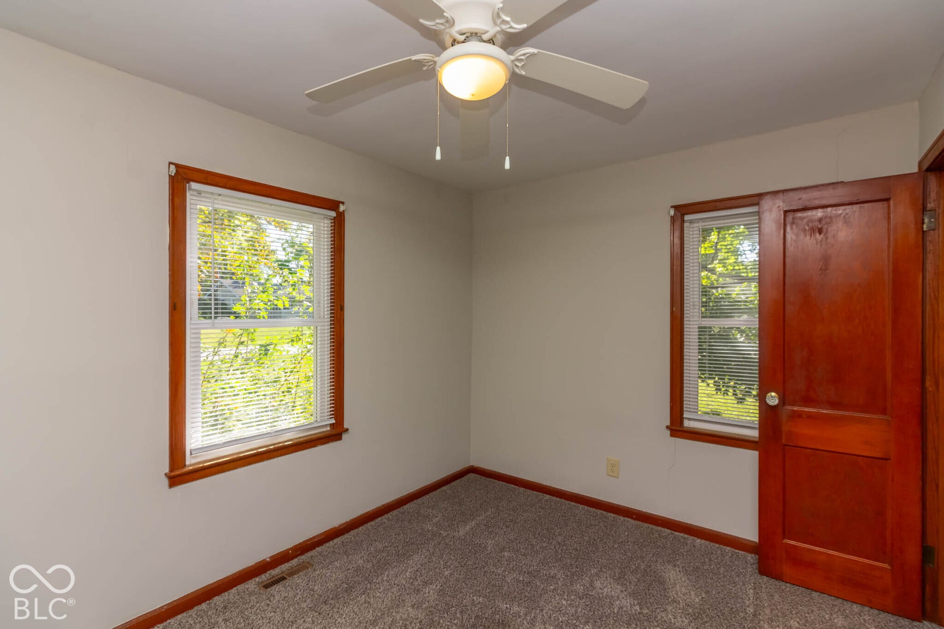 Neutral-toned interior space featuring a ceiling-mounted fan with integrated lighting, wood-trim windows, and a solid wood door
