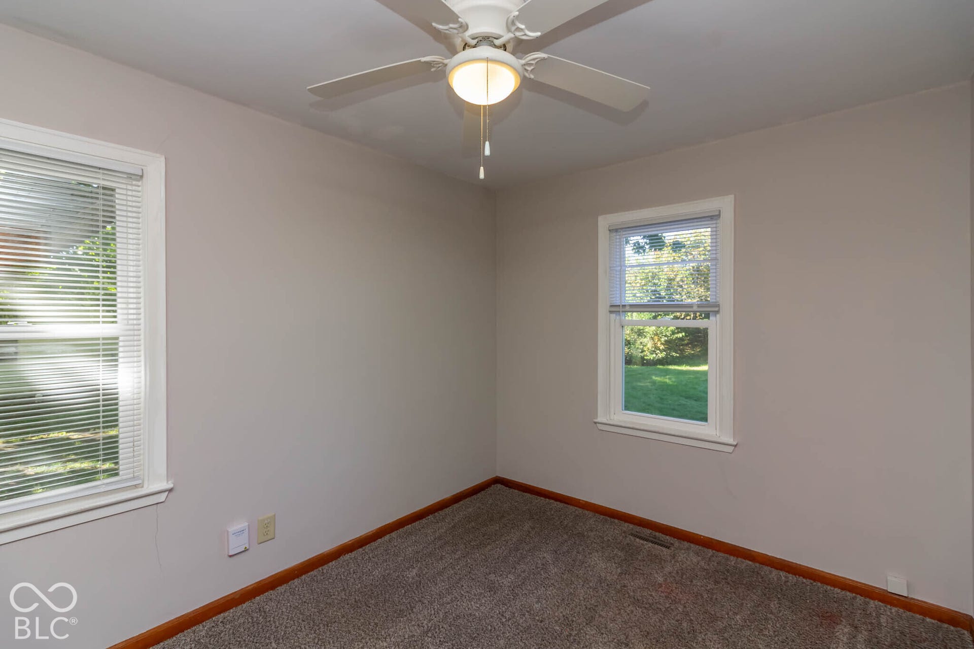 Neutral-toned interior space featuring carpeting, white-finish ceiling fan with integrated lighting, and dual windows with horizontal blinds