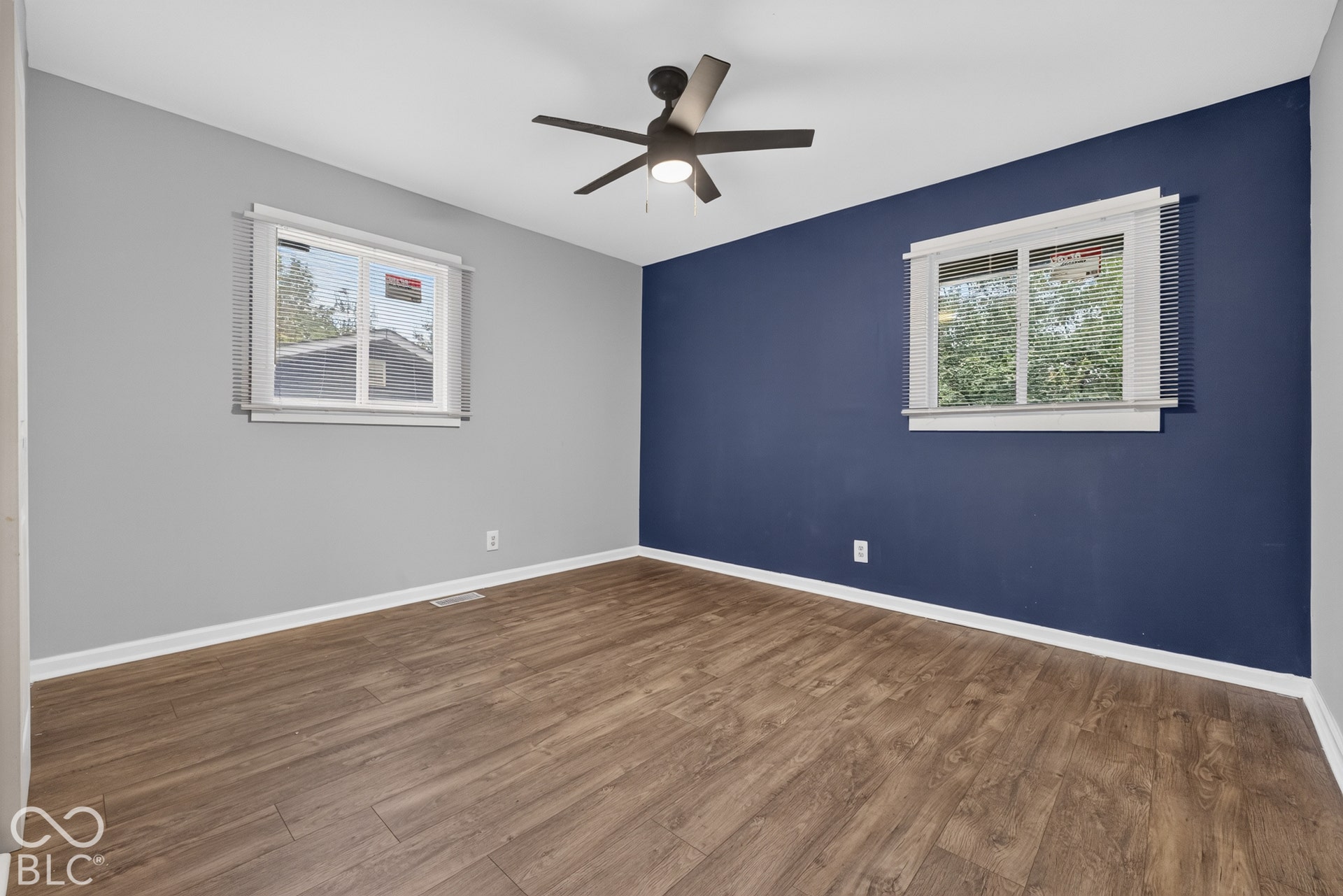 Room featuring wood-finish flooring, two windows with horizontal blinds, and a central ceiling fan with an integrated light fixture