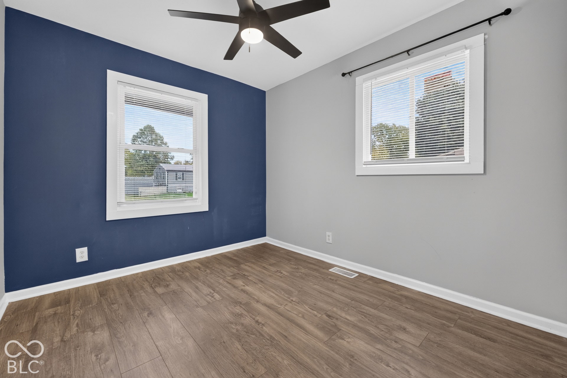 Room featuring wood-finish flooring, a dark-toned accent wall, a contrasting light grey wall, and a multi-blade ceiling fan with integrated lighting