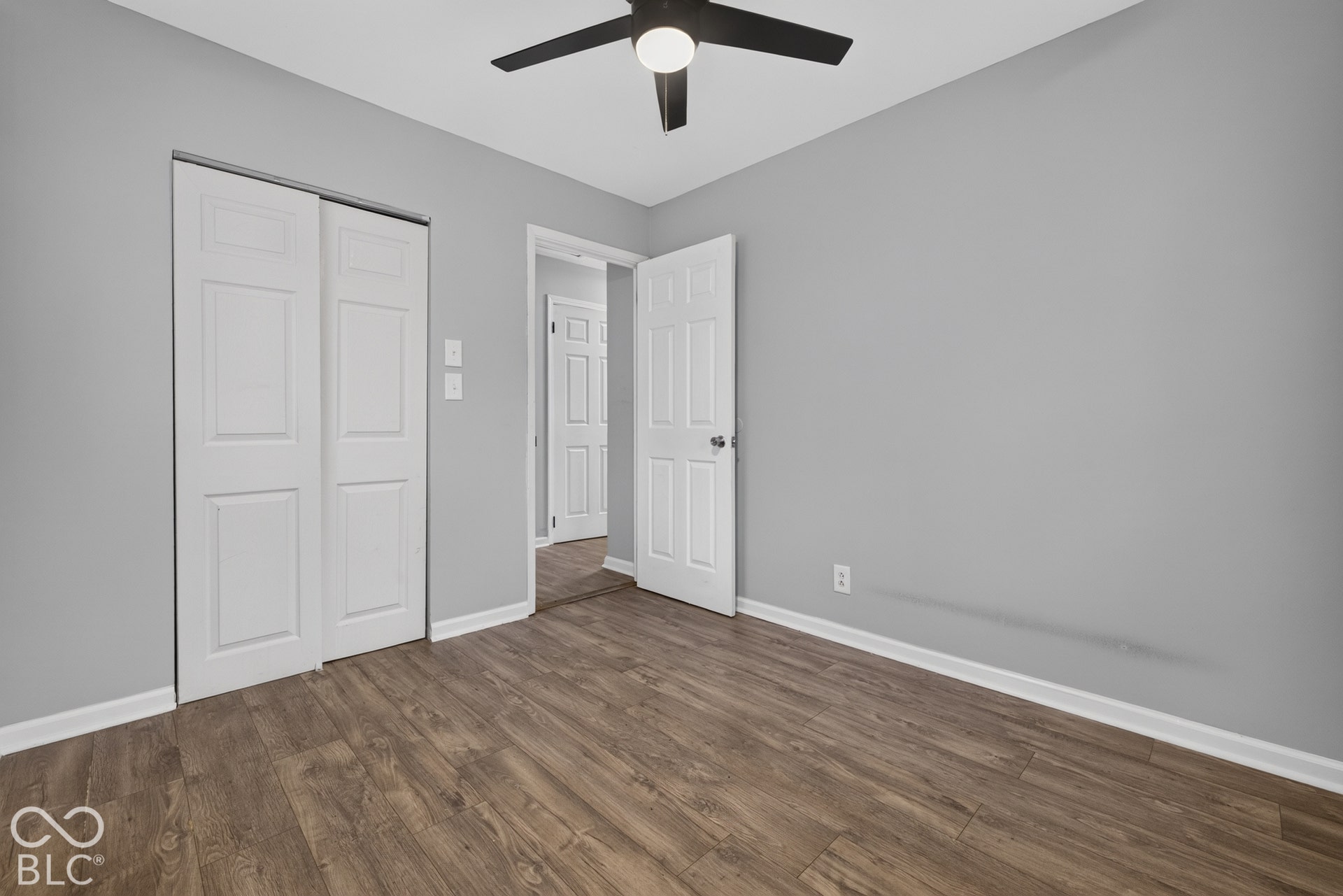 Wood-finish flooring, neutral gray wall paint, dual-panel closet doors, and a ceiling-mounted light fixture with fan blades