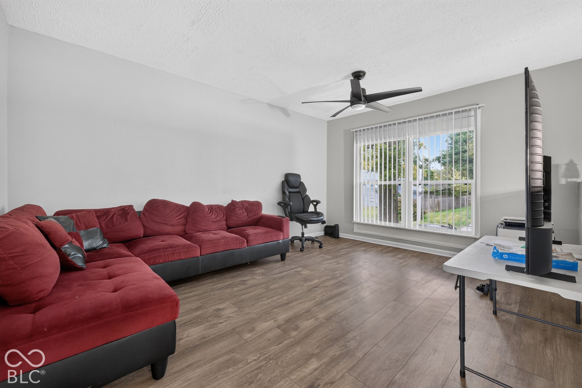 Wood-finish flooring, matte black ceiling fan, and large windows with vertical blinds