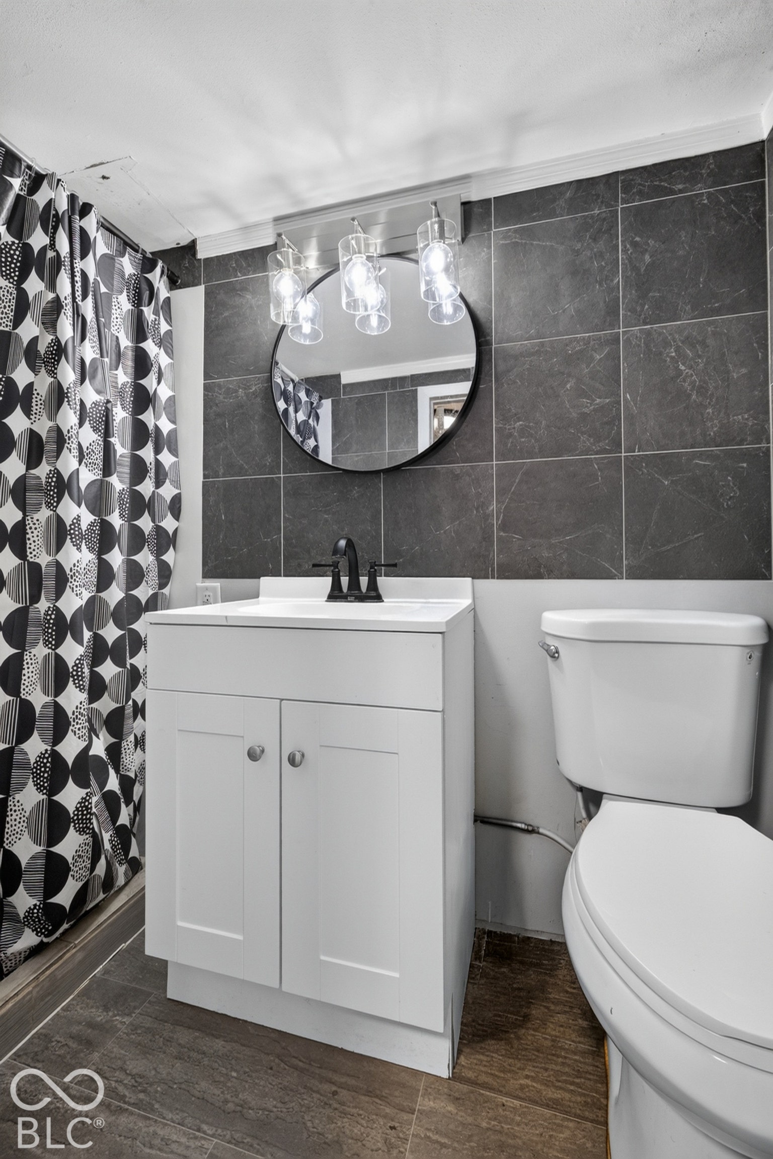 White shaker-style vanity with a solid-surface countertop, dark matte faucet, circular wall-mounted mirror, three-bulb vanity lighting fixture, and large-format dark tile backsplash