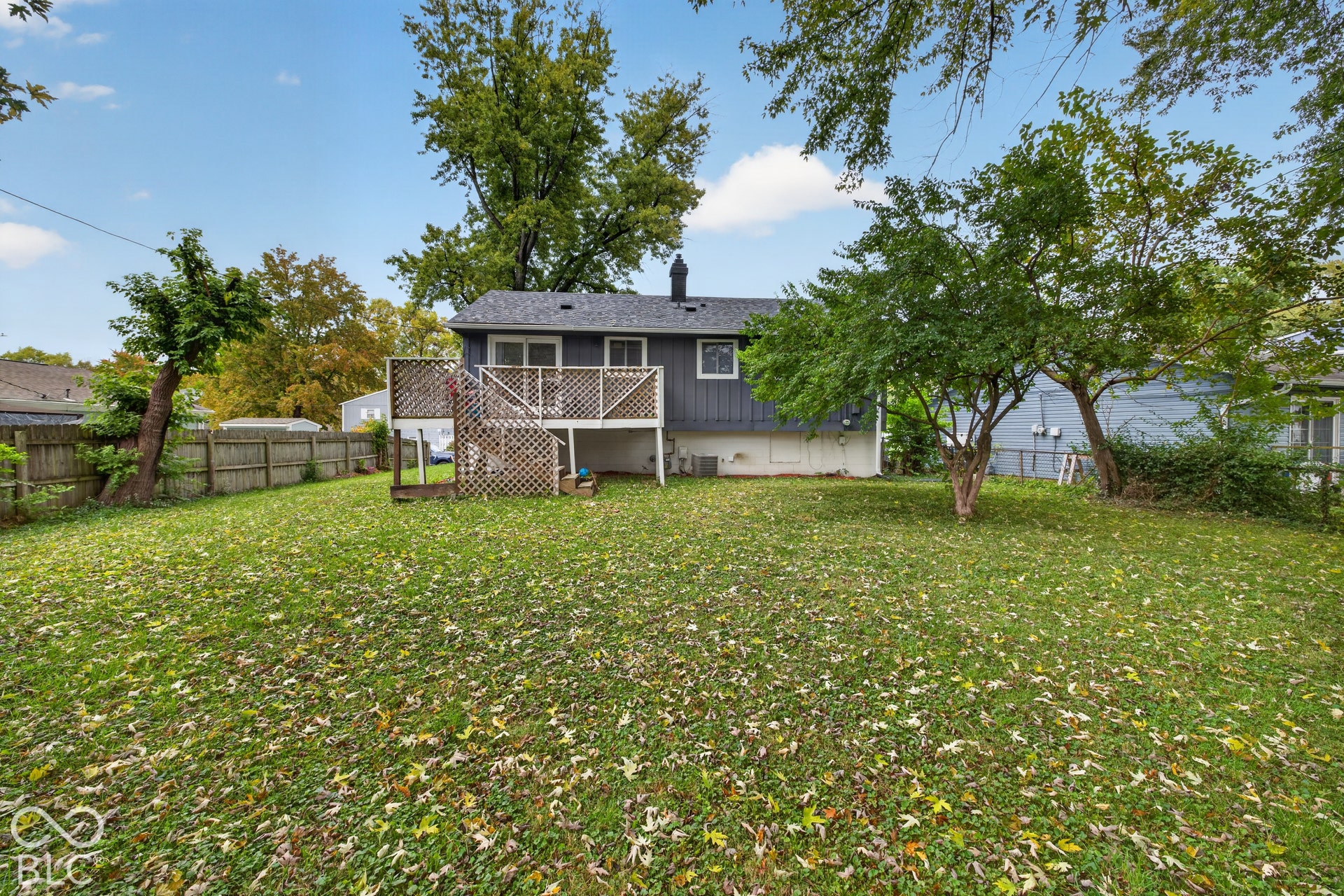 Rear exterior featuring a raised wooden deck with lattice privacy screening, expansive grass lawn, mature trees, and perimeter wood fencing