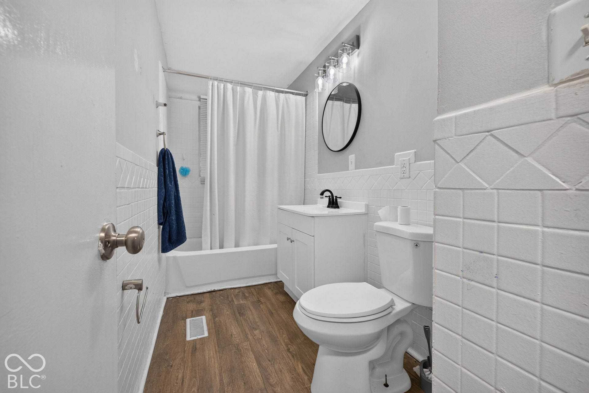 Bathroom featuring white tile wainscoting with decorative inlay, wood-finish flooring, white vanity with a single basin, matte black faucet, and a round wall-mounted mirror