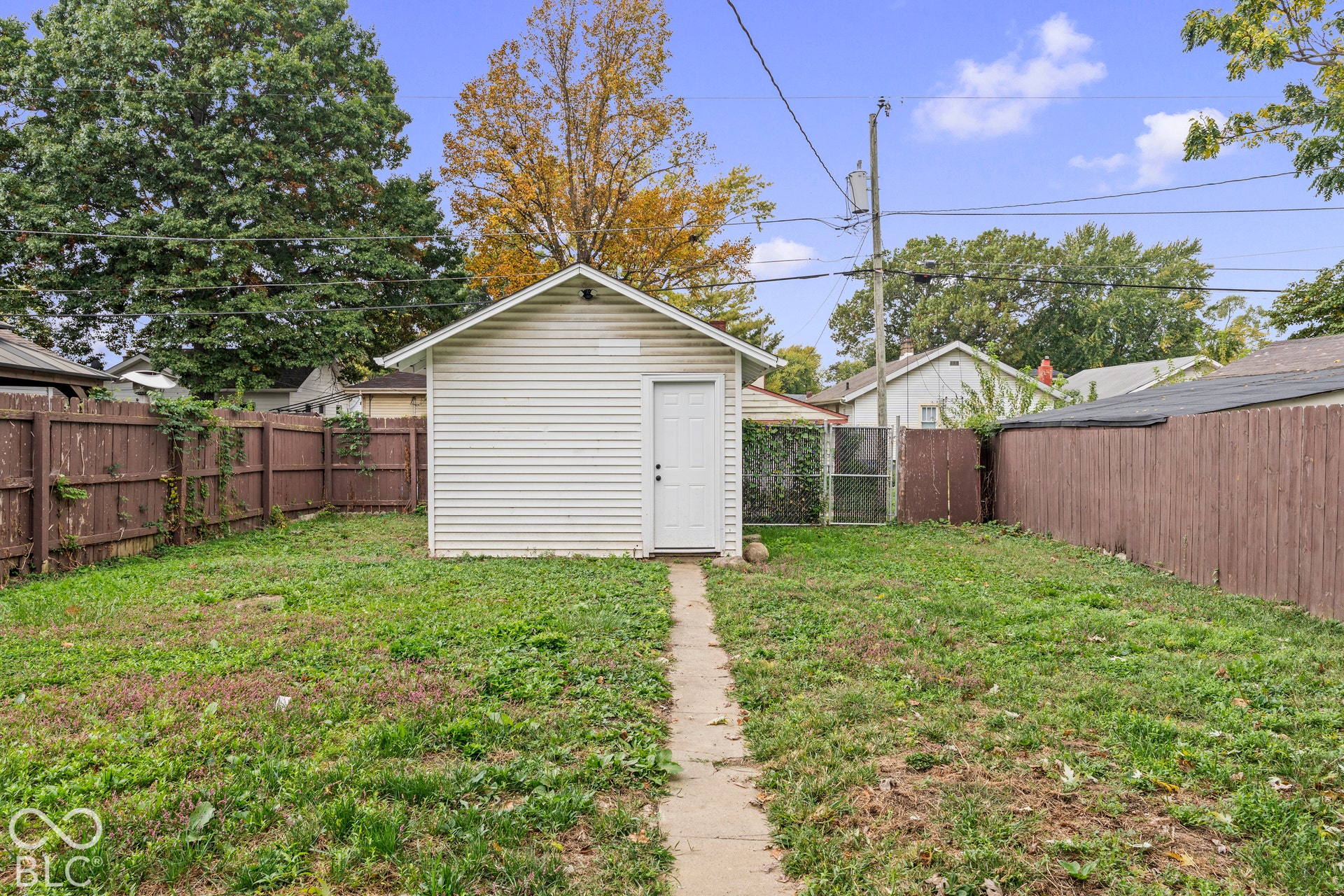Detached exterior structure with white horizontal siding and a single entry door, situated on a lot enclosed by wood panel fencing, featuring a concrete pathway