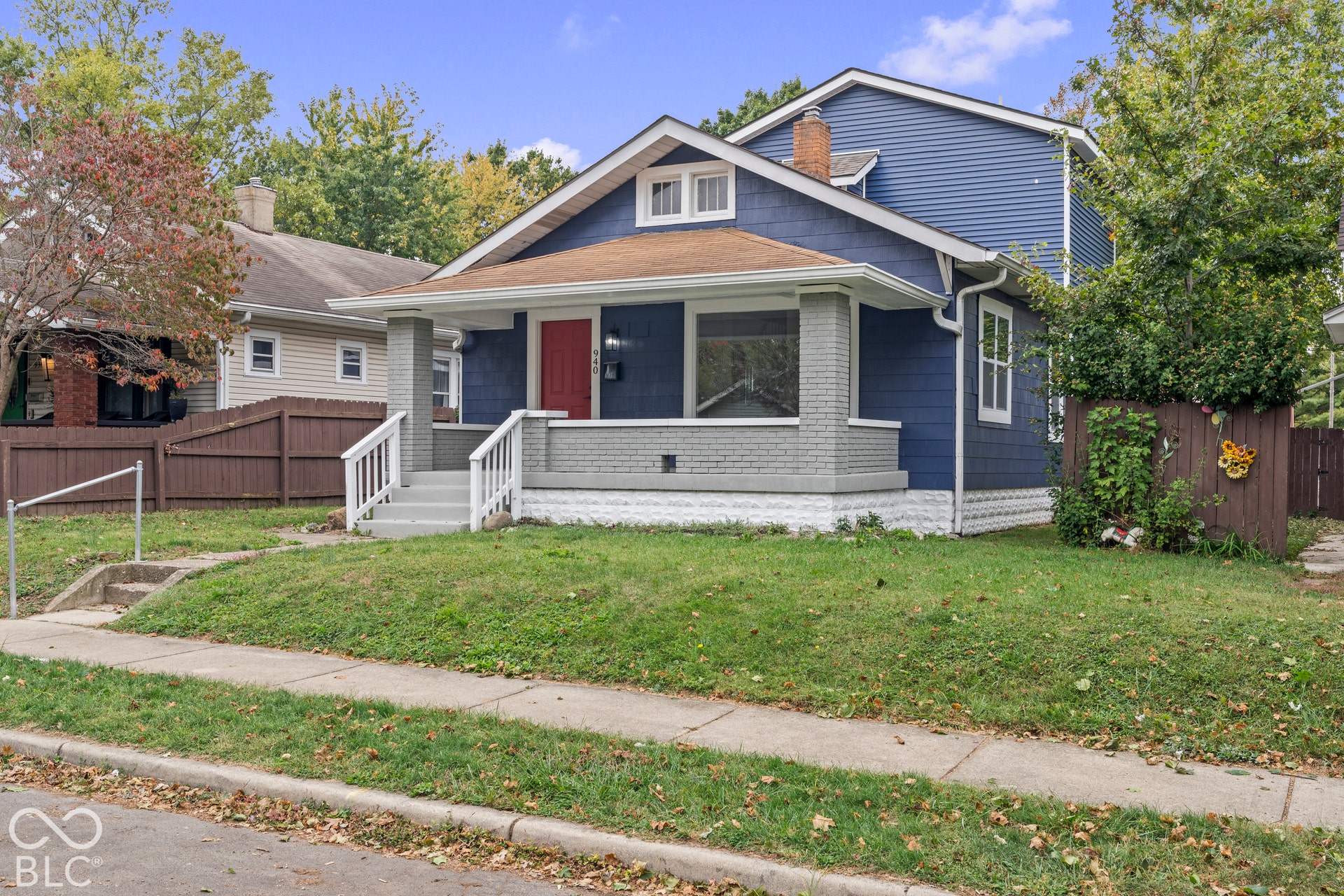 Craftsman-style exterior featuring blue horizontal siding, a covered front porch with painted brick columns, a prominent chimney, and wooden fencing