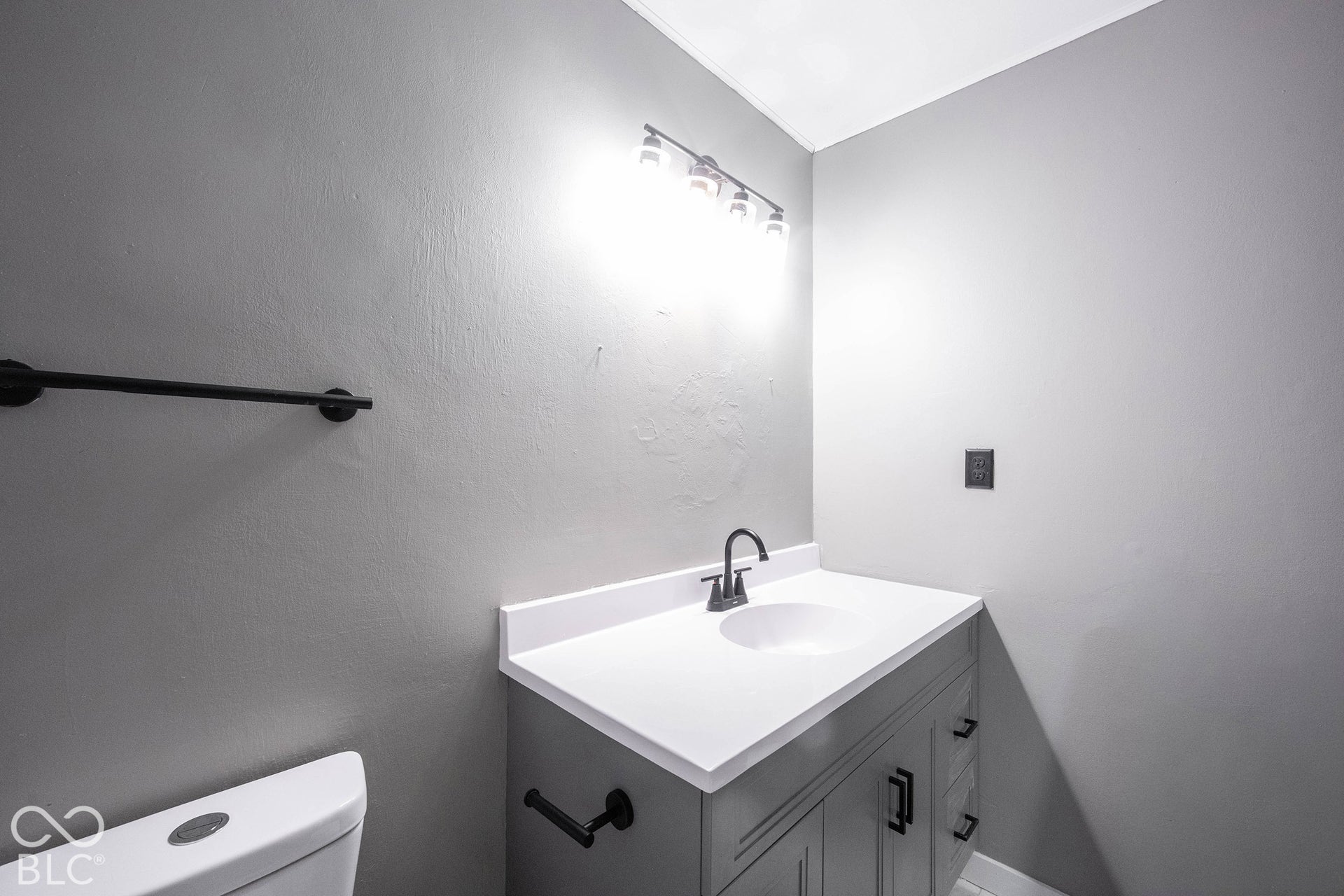 Vanity with white solid-surface countertop and integrated sink, matte black gooseneck faucet, shaker-style cabinetry, matte black hardware, and four-light vanity fixture