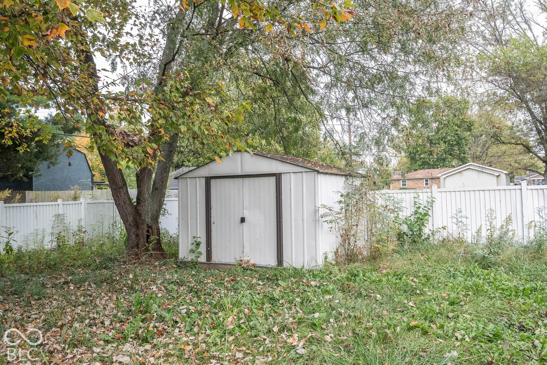 Exterior storage structure featuring metal siding, double doors, and a gable roof, enclosed by a white privacy fence