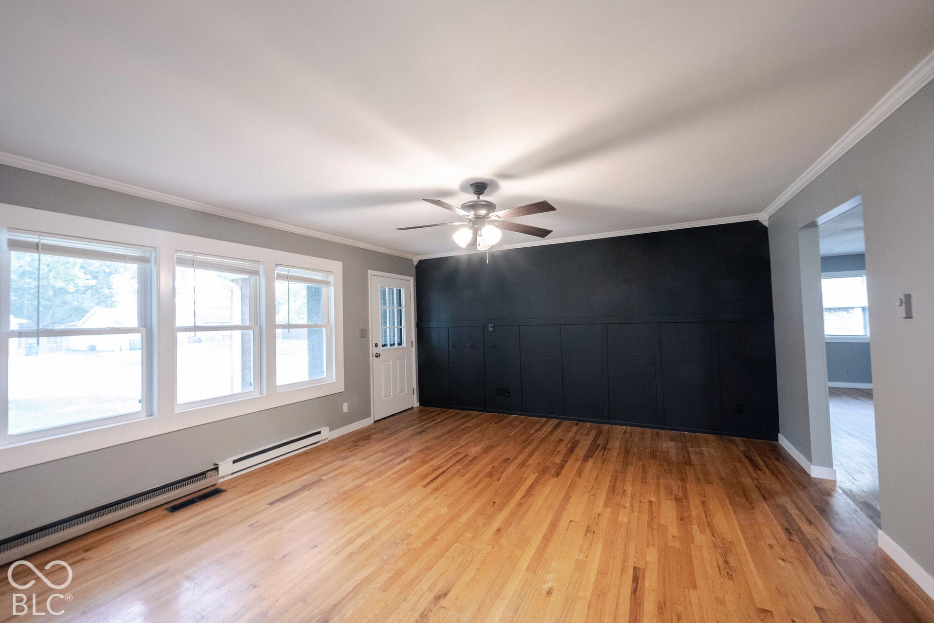 Wood-finish flooring, accent wall with paneled wainscoting, three-light ceiling fan, and large windows with white trim