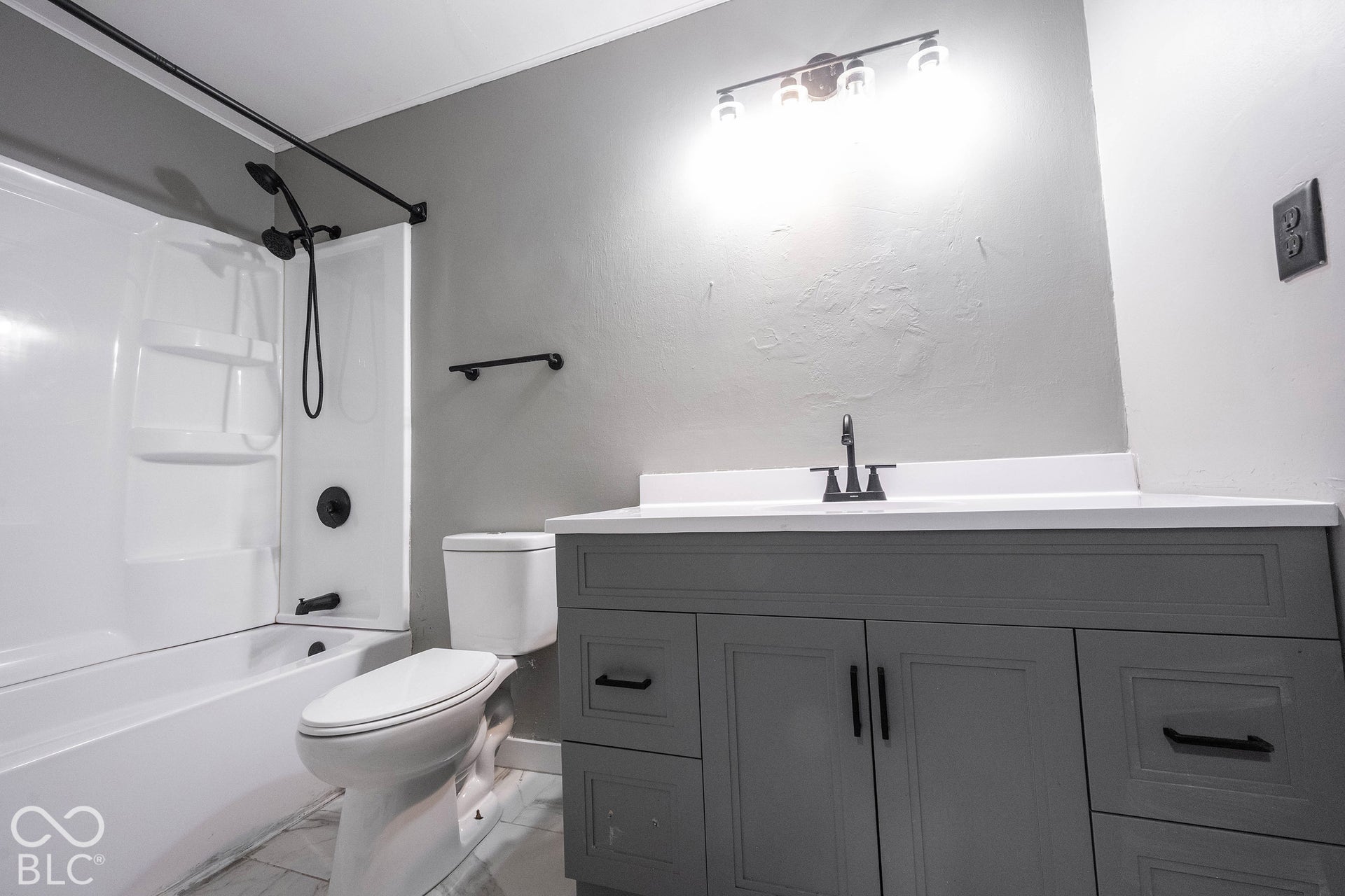 Bathroom featuring a gray vanity with shaker-style cabinetry, white countertop with an integrated basin, matte black faucet and hardware, tub-shower combination with matching matte black fixtures, and a four-bulb vanity light