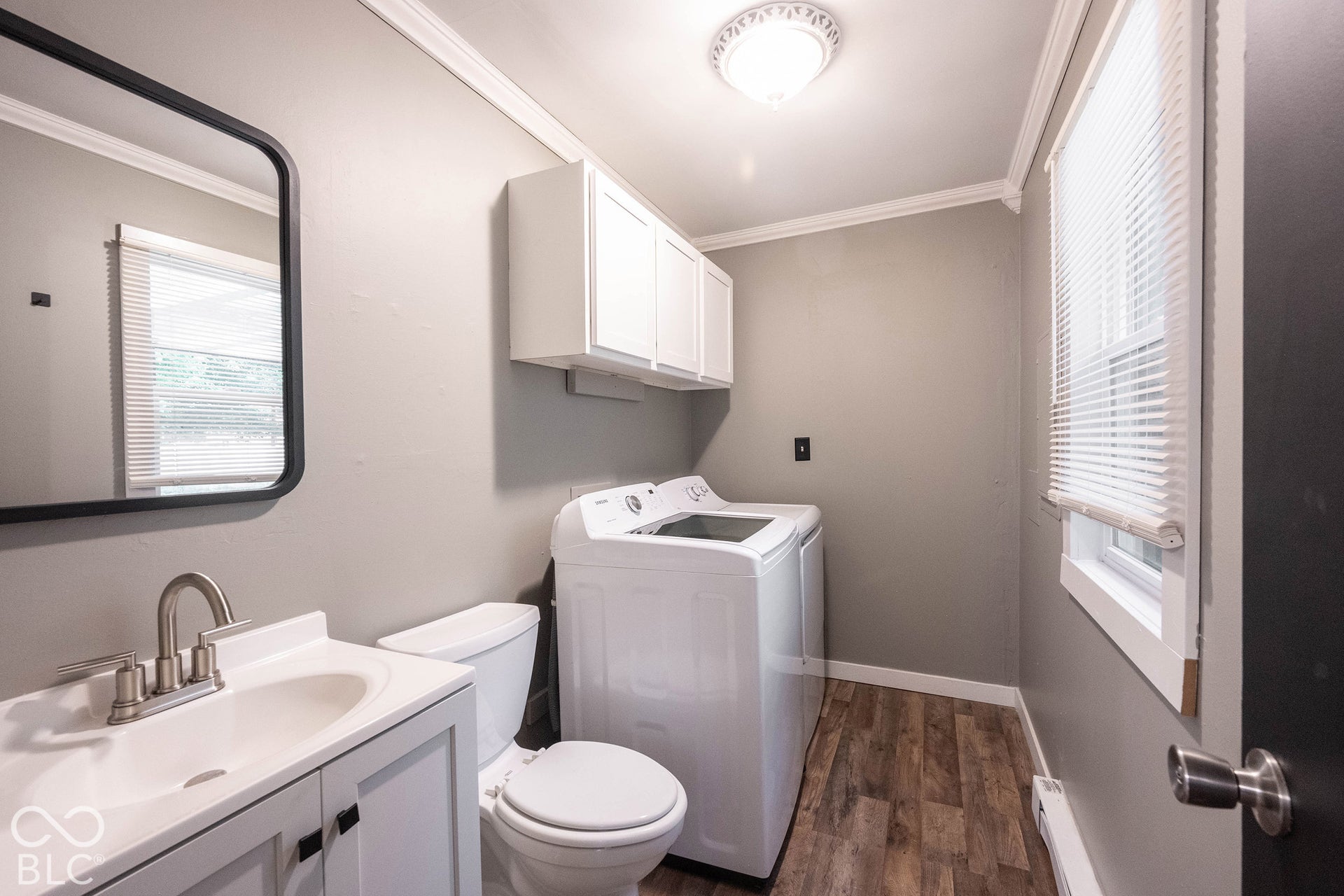 Utility space featuring wood-finish flooring, white wall-mounted cabinetry, a vanity with a drop-in sink and metallic hardware, and a window with integrated blinds