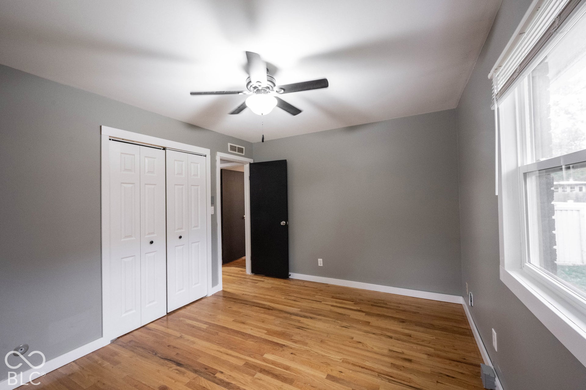 Wood-finish flooring, dual-panel closet doors, ceiling fan with integrated lighting, and white window trim