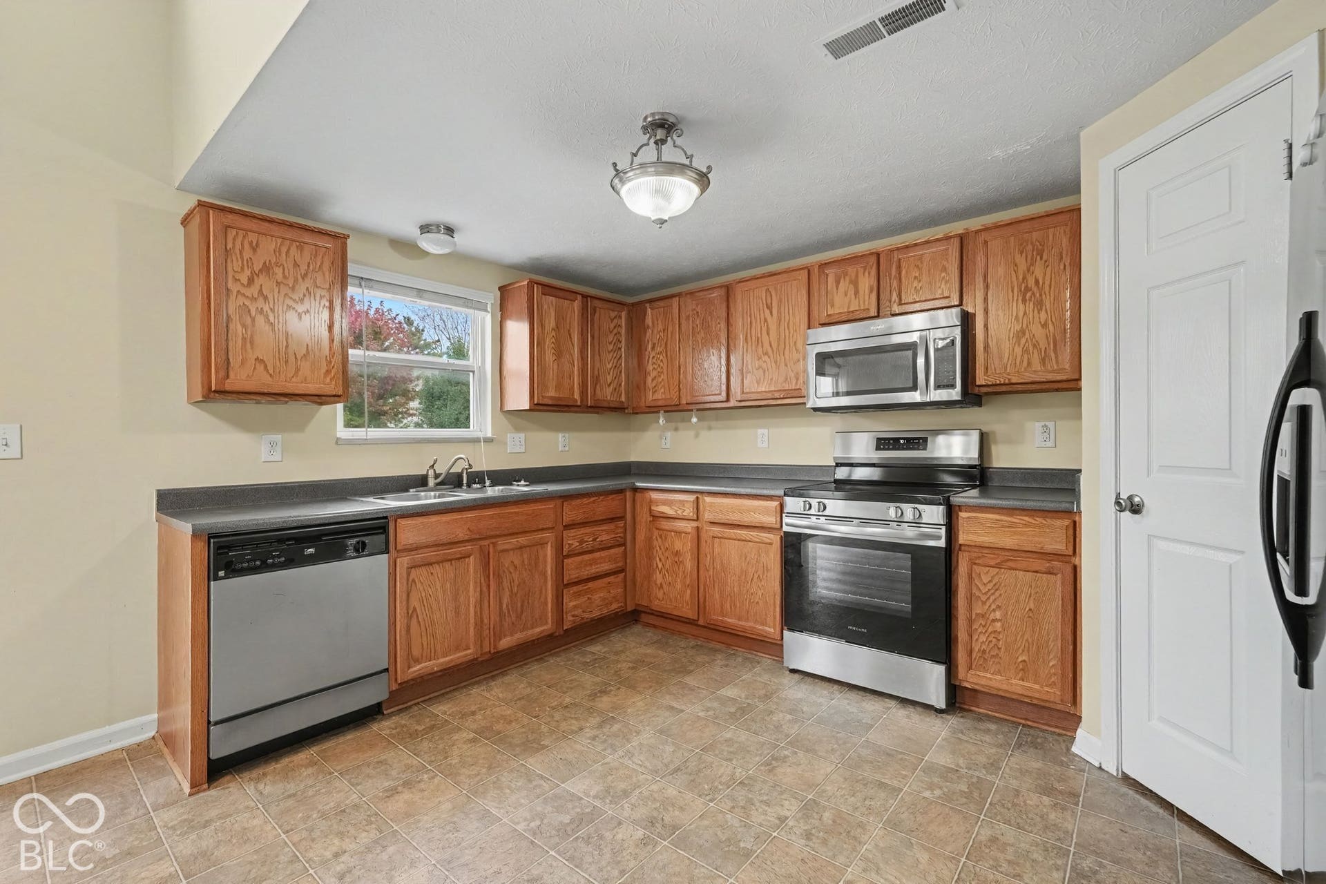 Kitchen featuring natural wood cabinetry, stainless steel appliances, dark solid-surface countertops, tile flooring, and a flush-mount light fixture