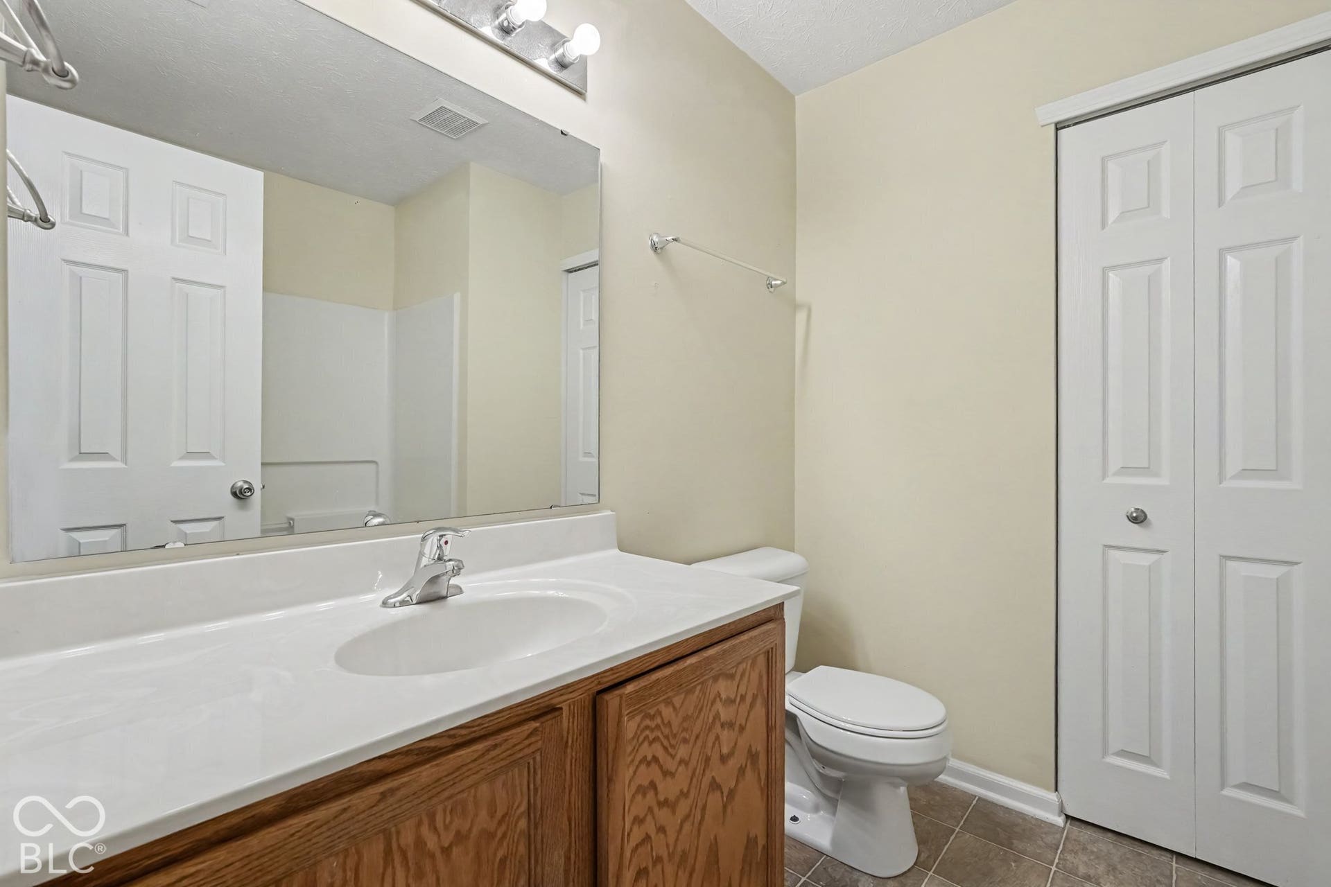 Bathroom vanity with wood-finish cabinetry, solid-surface integrated sink, expansive wall-mounted mirror, brushed-finish hardware, and bifold closet doors