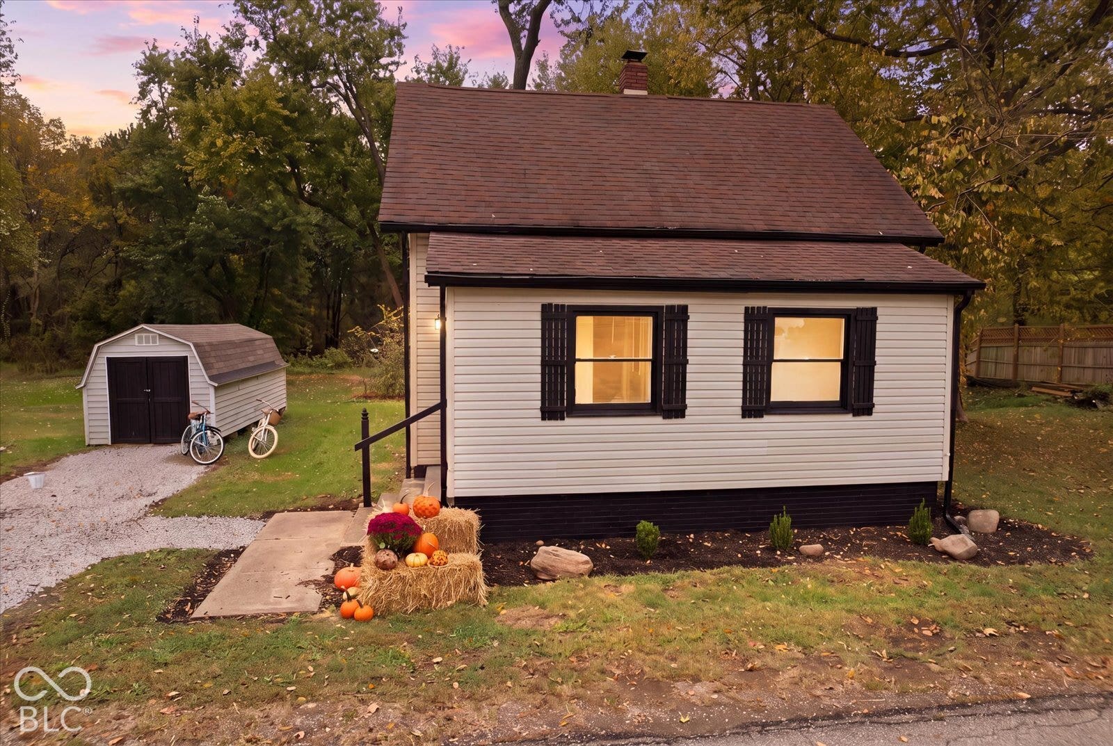 Virtually Altered - Single-story residence featuring light-colored siding, black shutter accents, a dark-toned roof, and a detached storage structure on a gravel drive