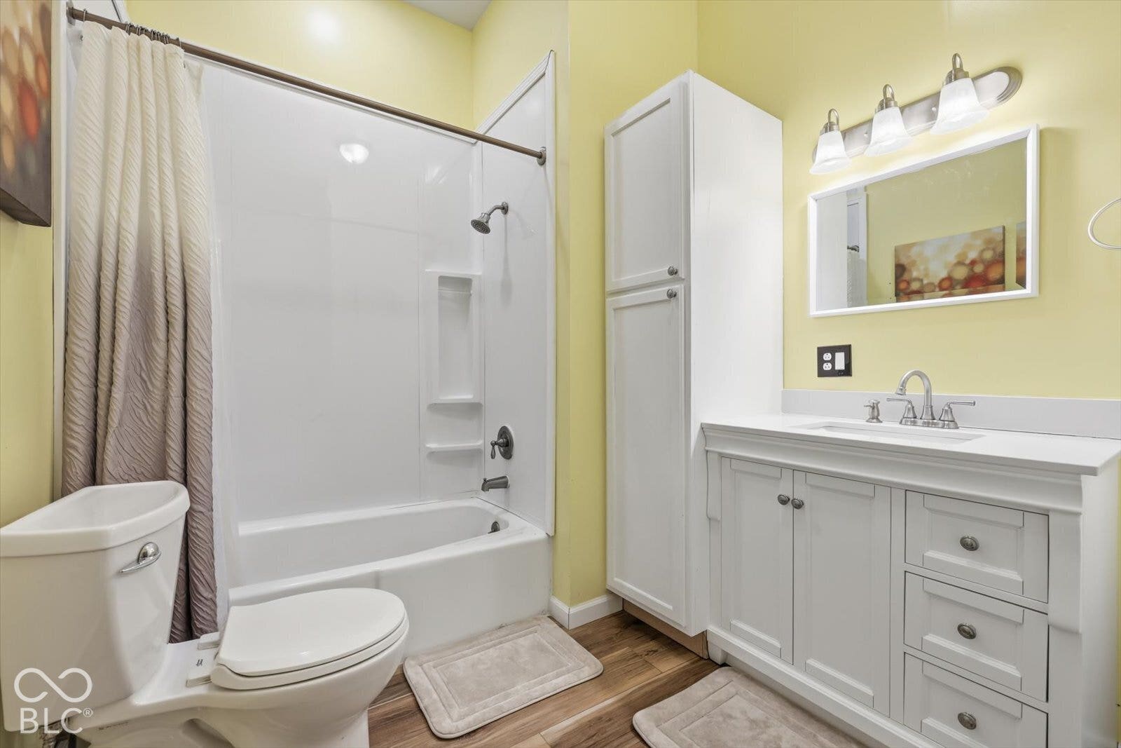 Bathroom featuring a white vanity with integrated cabinetry, solid surface countertop with undermount sink, tub and shower combination with molded shelving, and wood-finish flooring