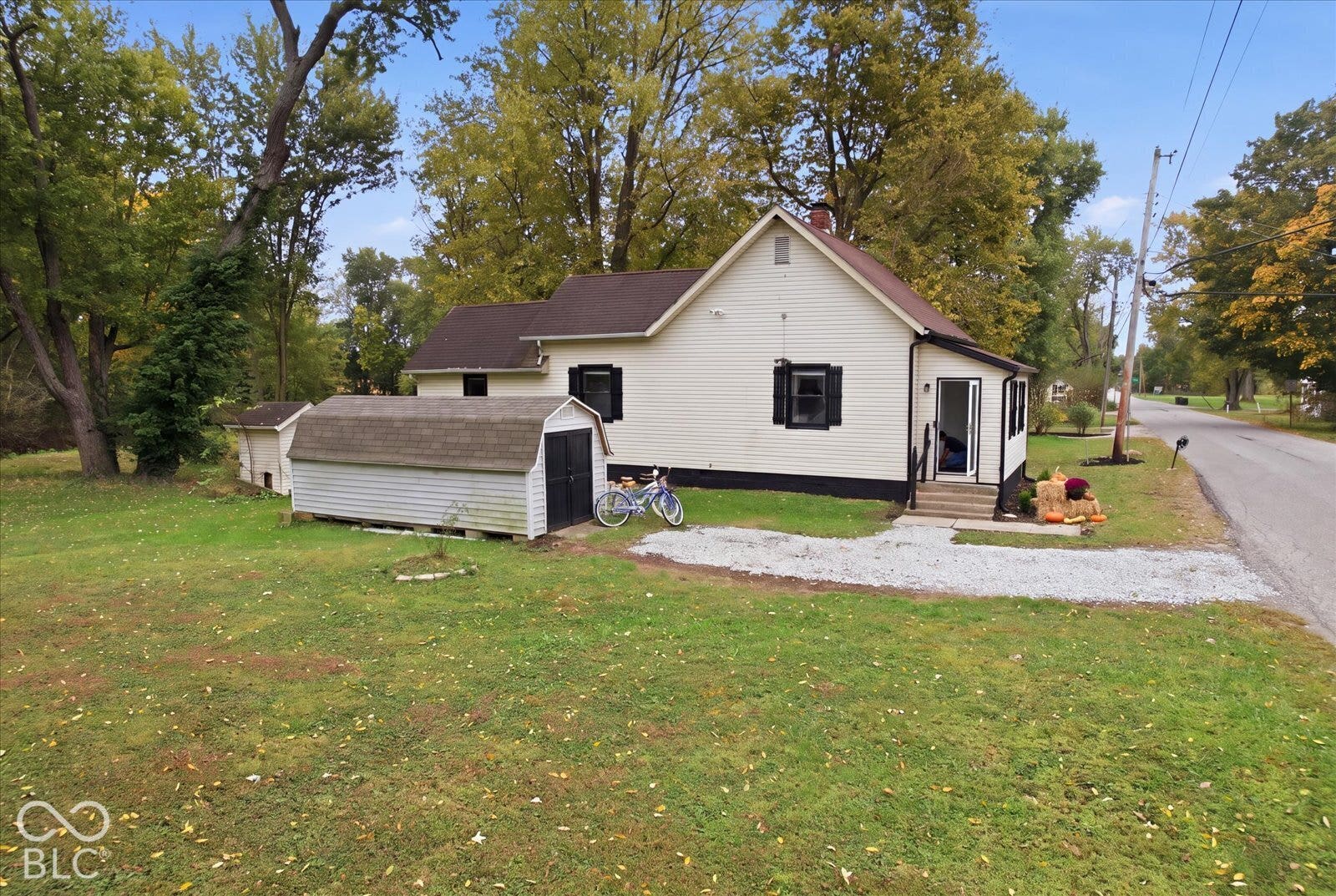 Single-story residence featuring light-toned horizontal siding, a pitched roof with dark shingles, black exterior shutters, a gravel driveway, and a detached storage structure