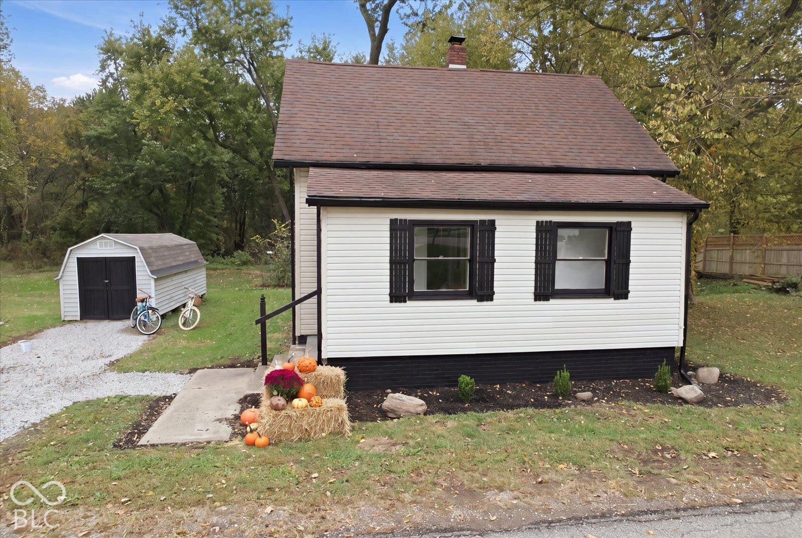 Single-story residence featuring light-colored horizontal siding, dark decorative shutters, a pitched shingle roof, chimney, and a detached outbuilding with a gravel driveway