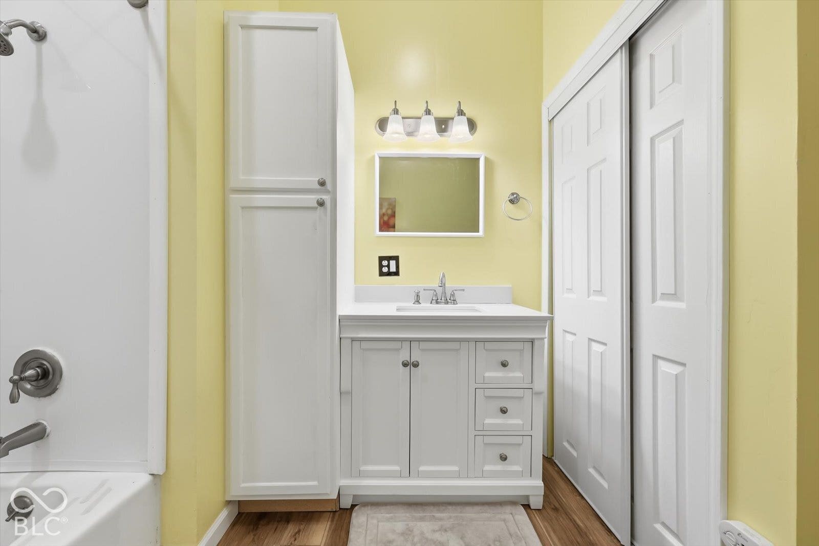 Bathroom vanity featuring a white finish, stone-composite countertop, integrated sink, and brushed-metal fixtures, complemented by a tall built-in storage cabinet and wood-finish flooring