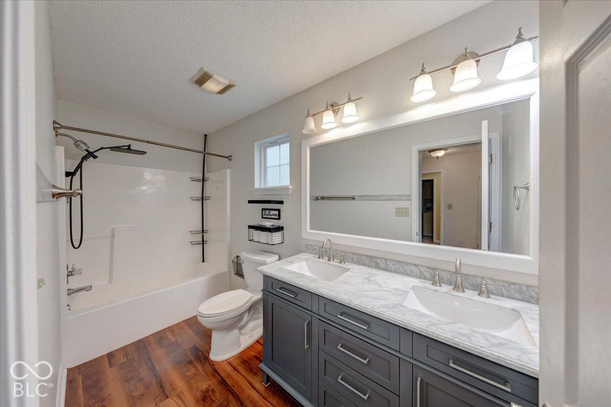 6405 Lonestar Drive, Indianapolis, IN 46237 Bathroom featuring a charcoal dual-sink vanity with a white countertop, wood-finish flooring, a full-width framed mirror, and a shower-tub combination with a matte black fixture