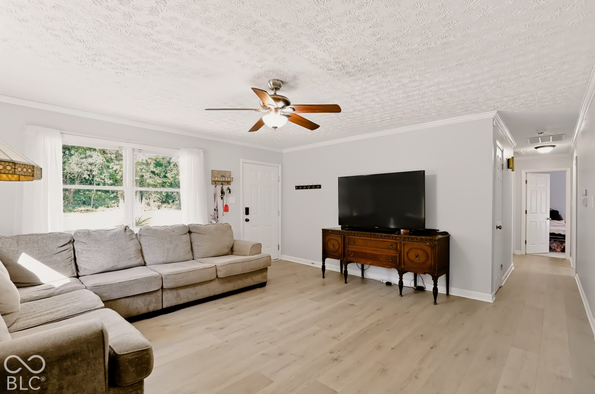 Living area featuring wood-finish flooring, a ceiling fan with integrated lighting, crown molding, and large windows