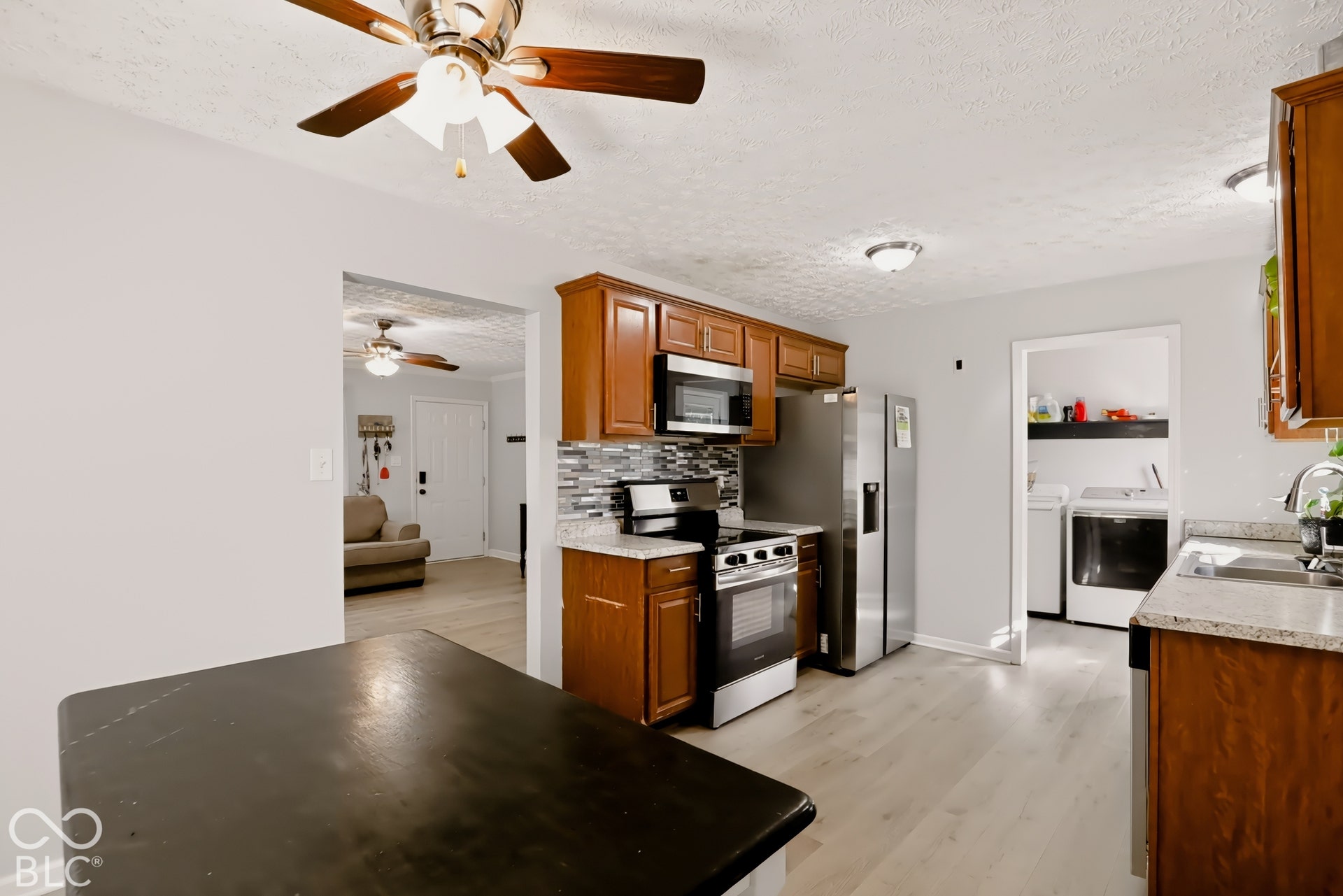 Kitchen featuring natural wood-finish cabinetry, stone-texture countertops, multi-tonal tile backsplash, stainless steel appliances, and wood-finish flooring